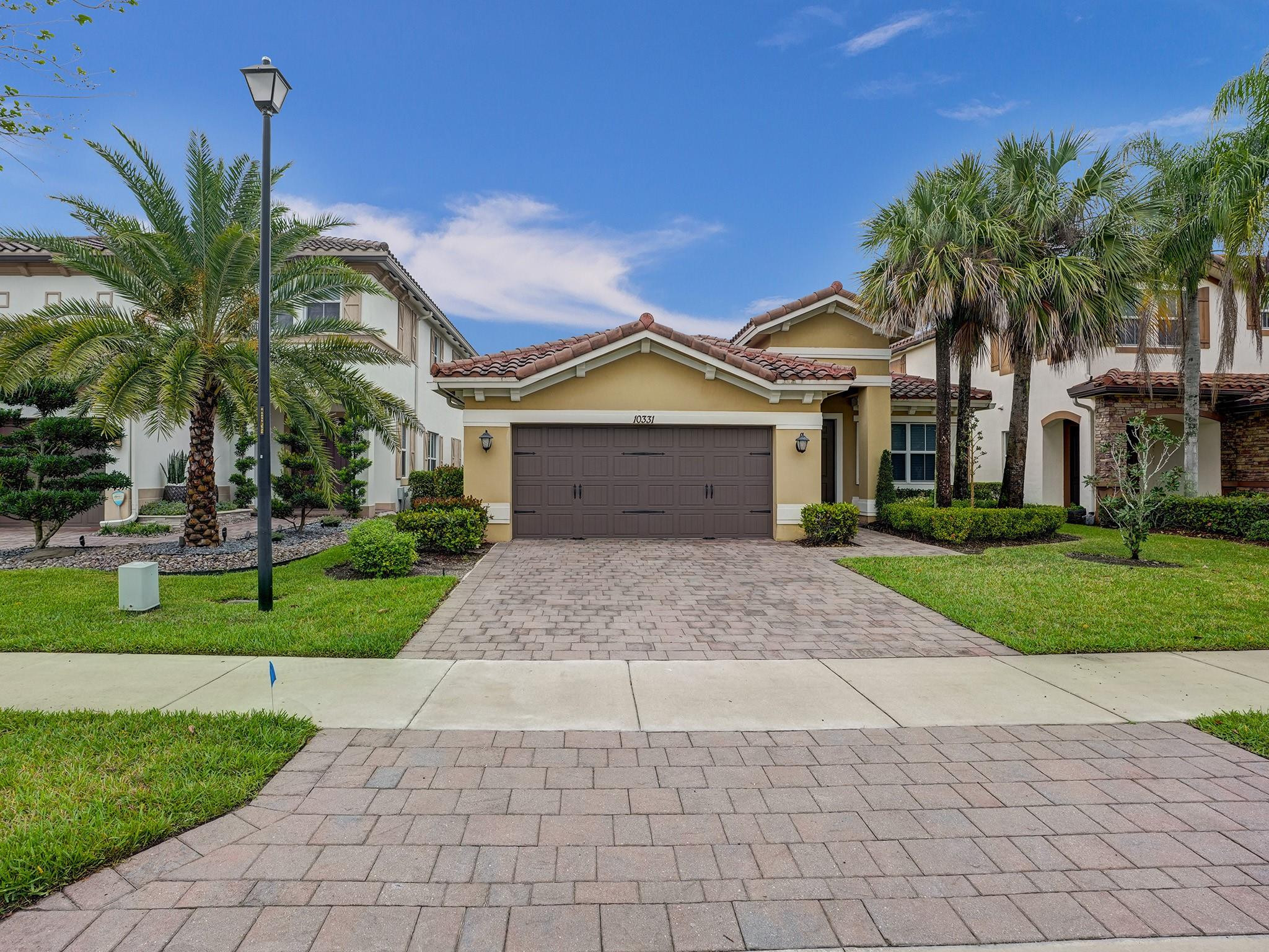 Tan stucco home with a double garage, terracotta tile roof, palm trees, and a broad paver driveway.