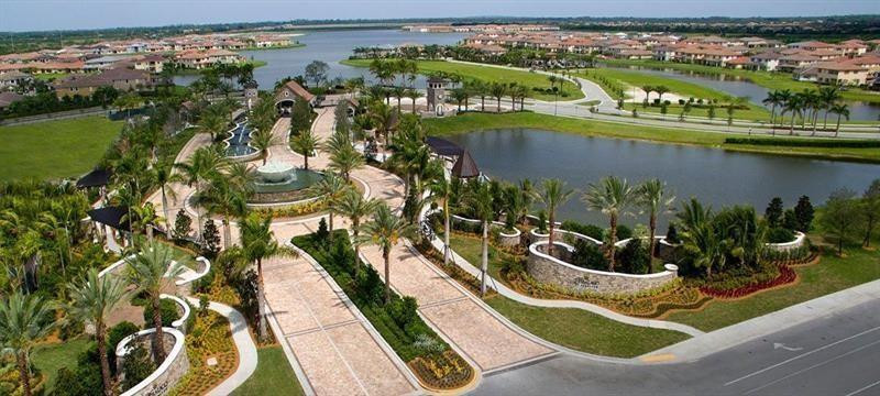 Aerial view of a landscaped residential entrance beside a lake, with palm-lined paths and homes.