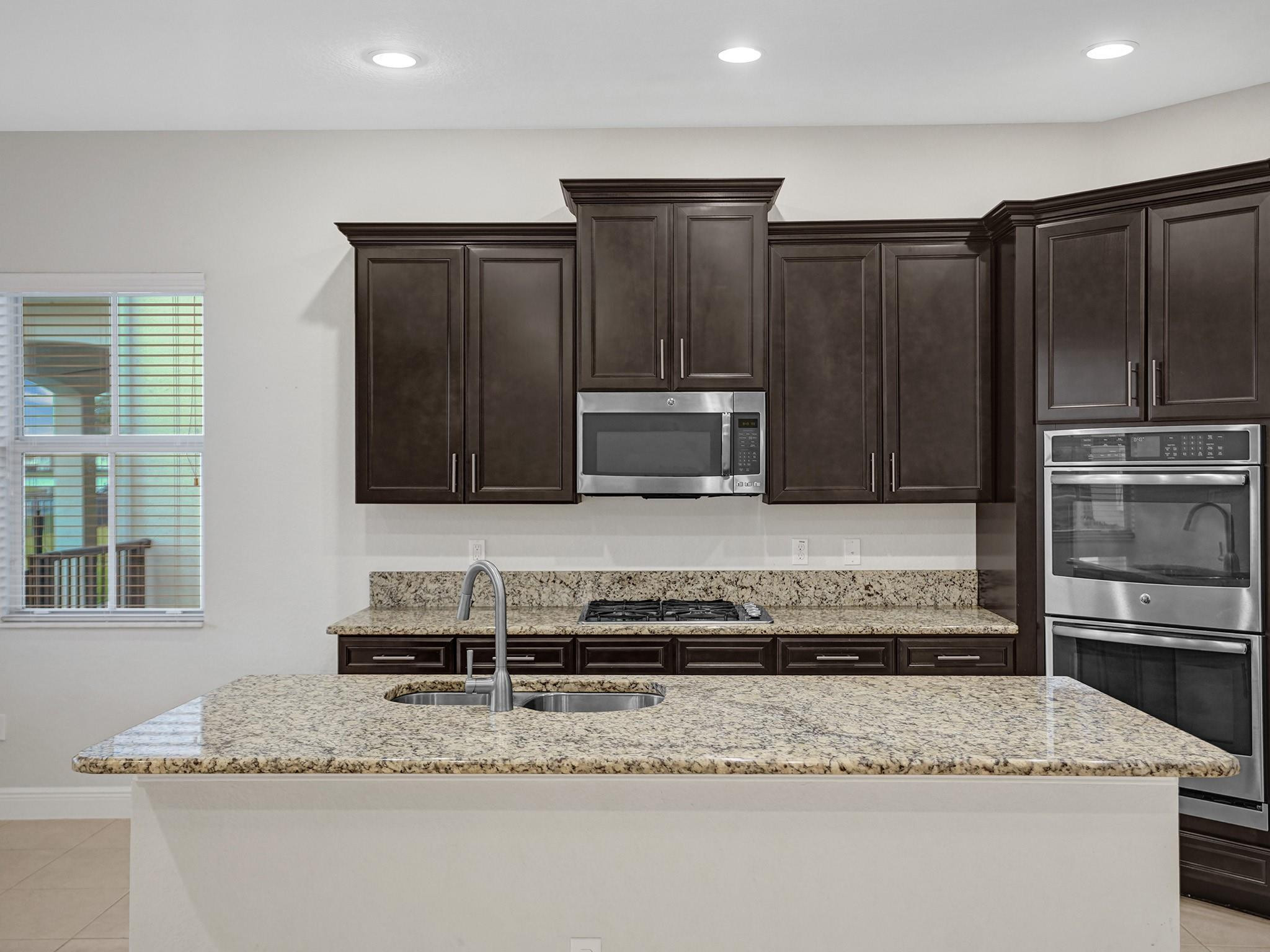 Kitchen with dark wood cabinets, granite island, double sink, and built-in stainless steel appliances.