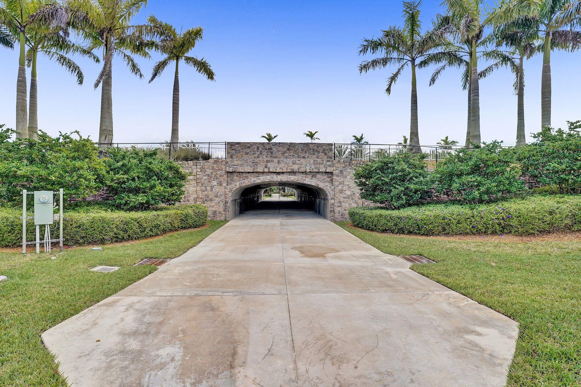 Landscaped driveway passing beneath a stone archway, framed by hedges and tall palm trees.
