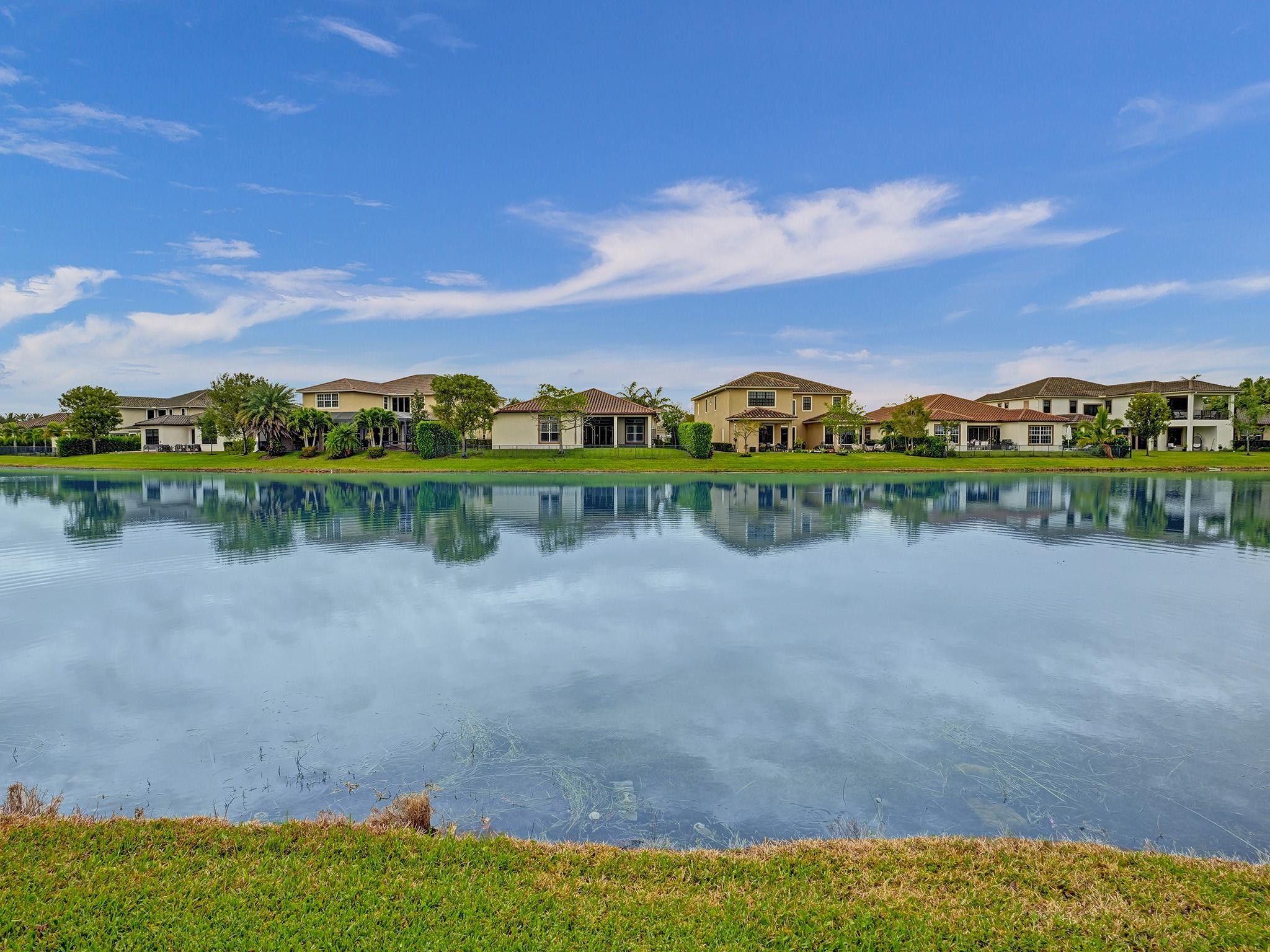 Lakefront homes reflected in calm water, viewed across a grassy bank.