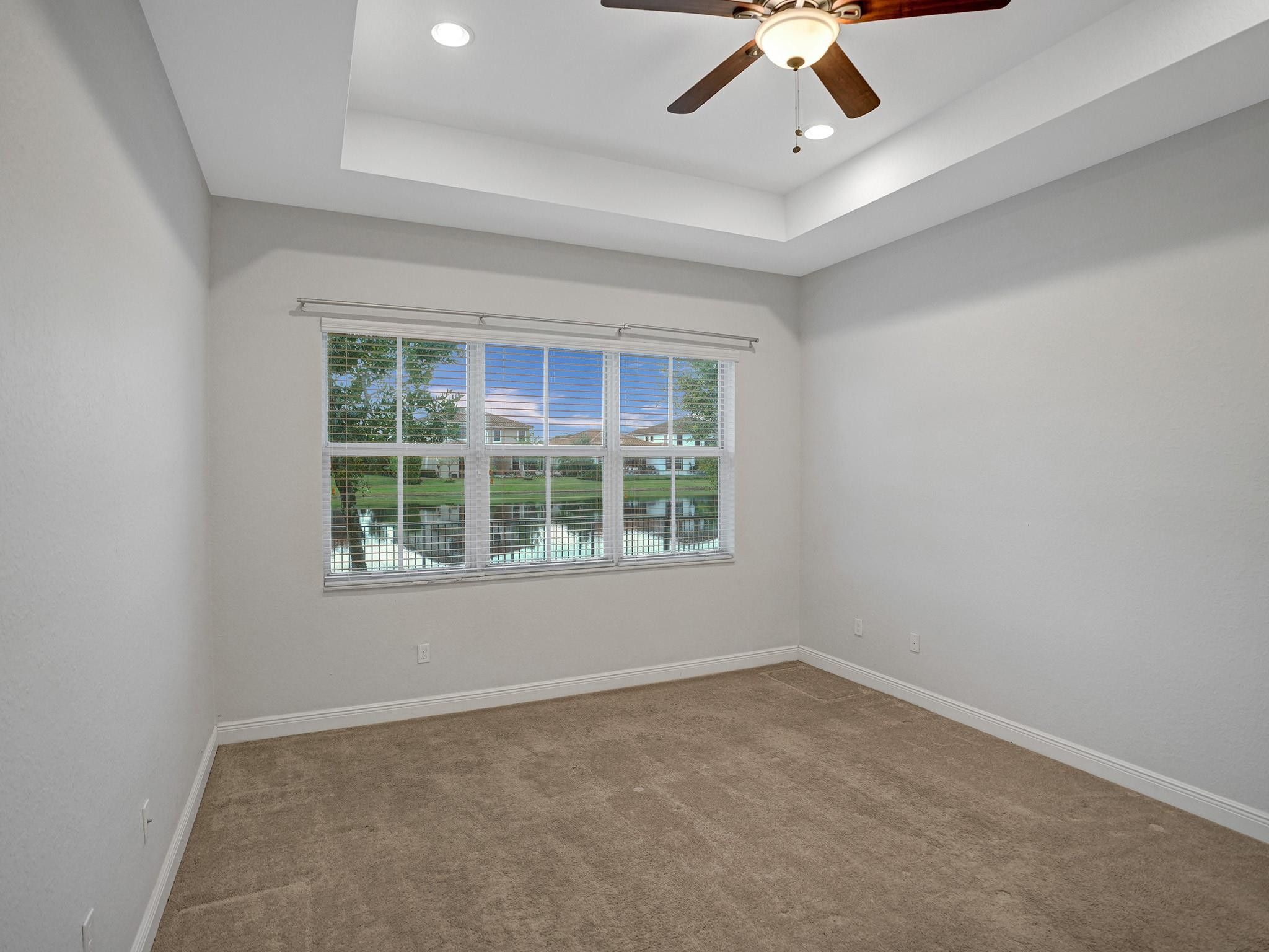 Empty carpeted bedroom with a tray ceiling and ceiling fan overlooking a pond.