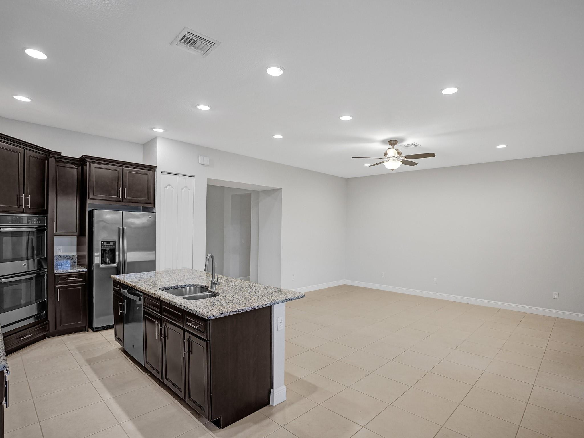 Kitchen with dark cabinets, stainless steel appliances, and a granite island opening to a large tiled room.
