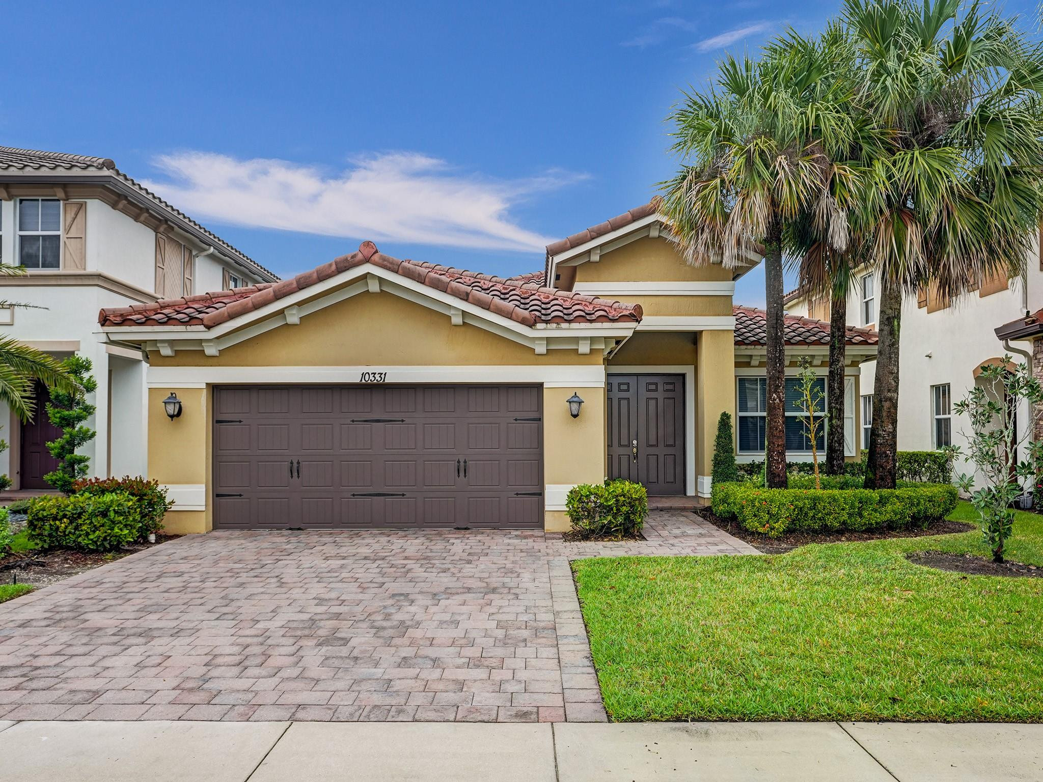 Yellow stucco home with a red tile roof, double garage, paver driveway, and palm trees.
