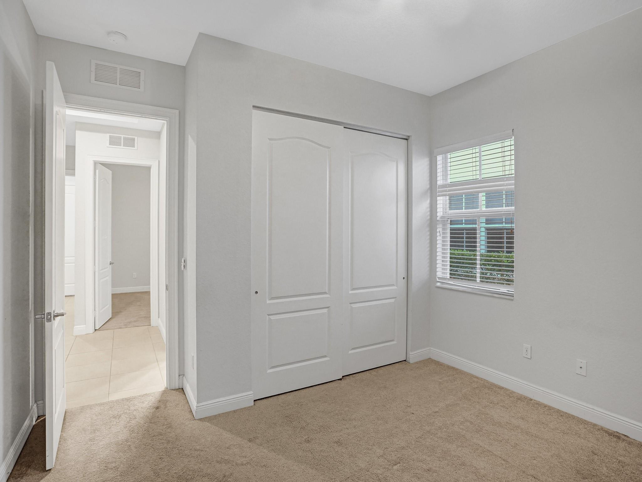 Empty bedroom with beige carpet, a large sliding-door closet, and a window with blinds.