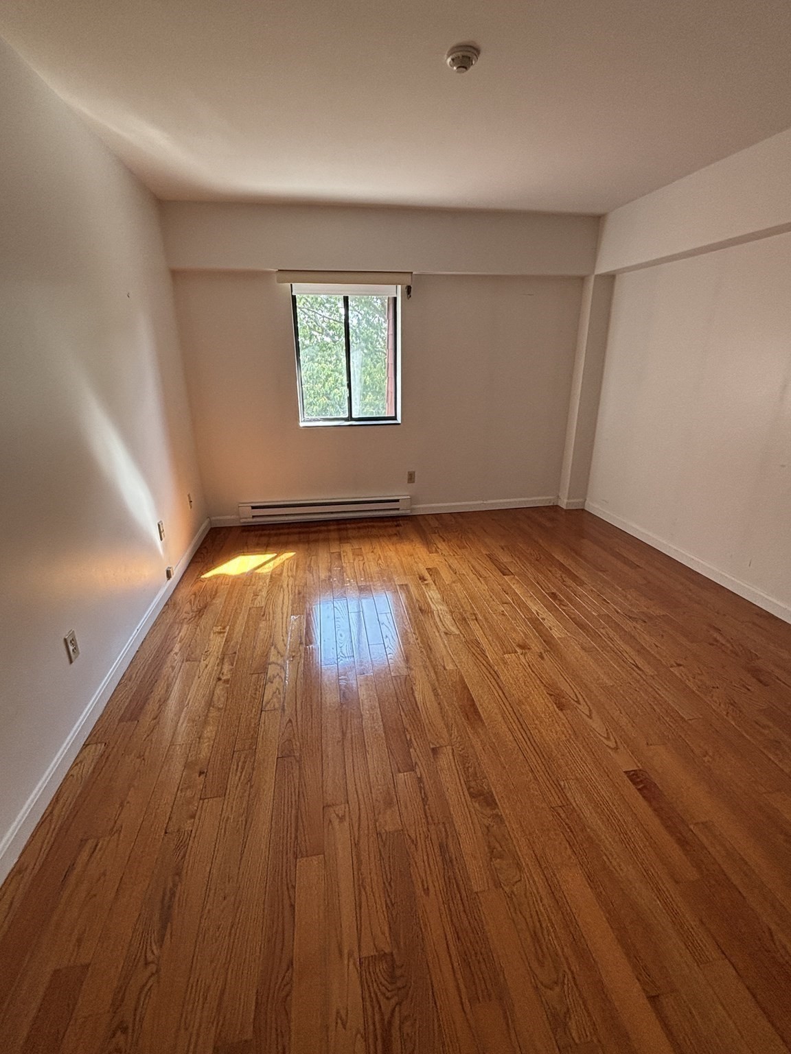 Empty room with polished hardwood floors, a window, and a baseboard heater.