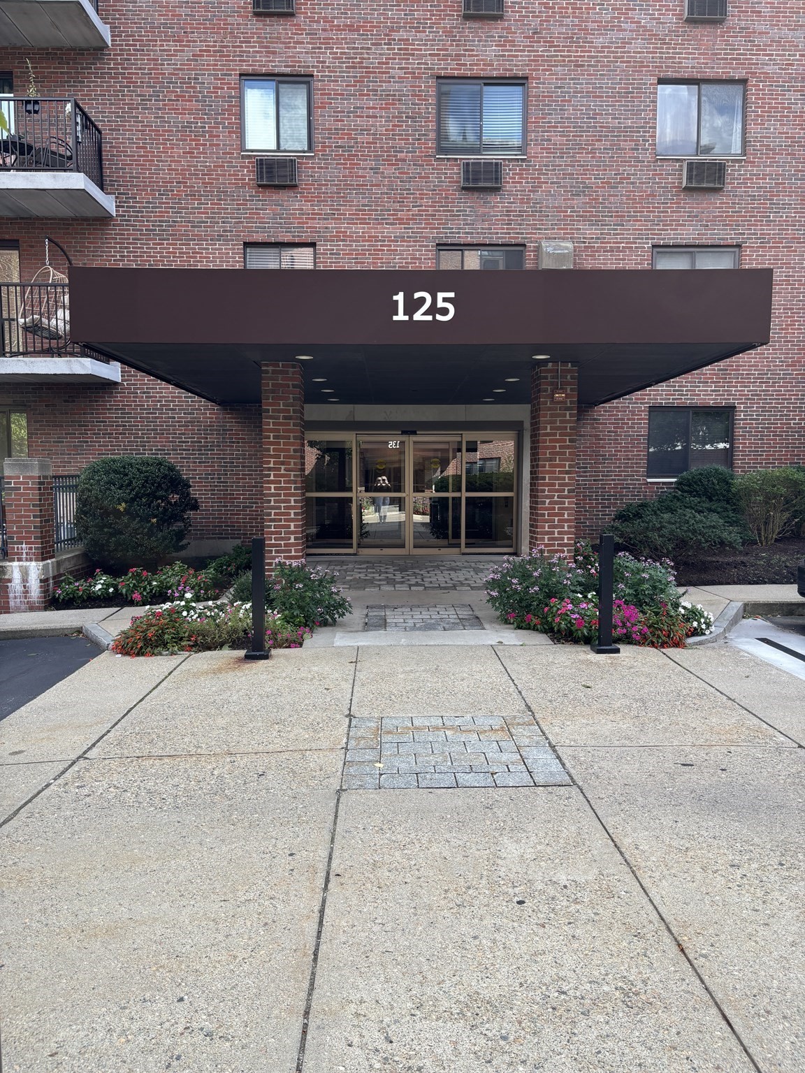 Brick apartment building entrance marked 125, with glass doors, balconies, and flower beds.