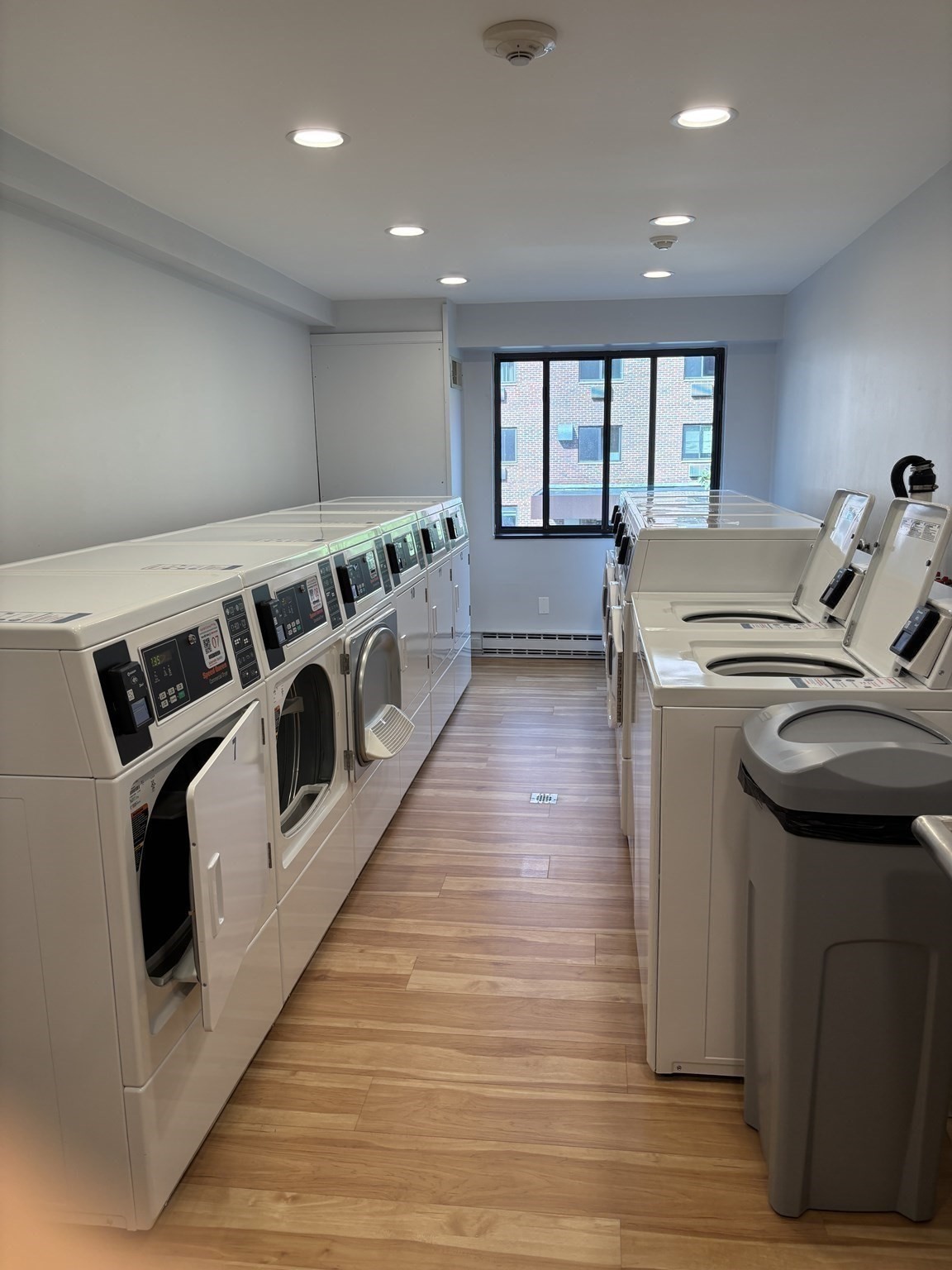 Shared laundry room with rows of commercial washers and dryers beside a large window.