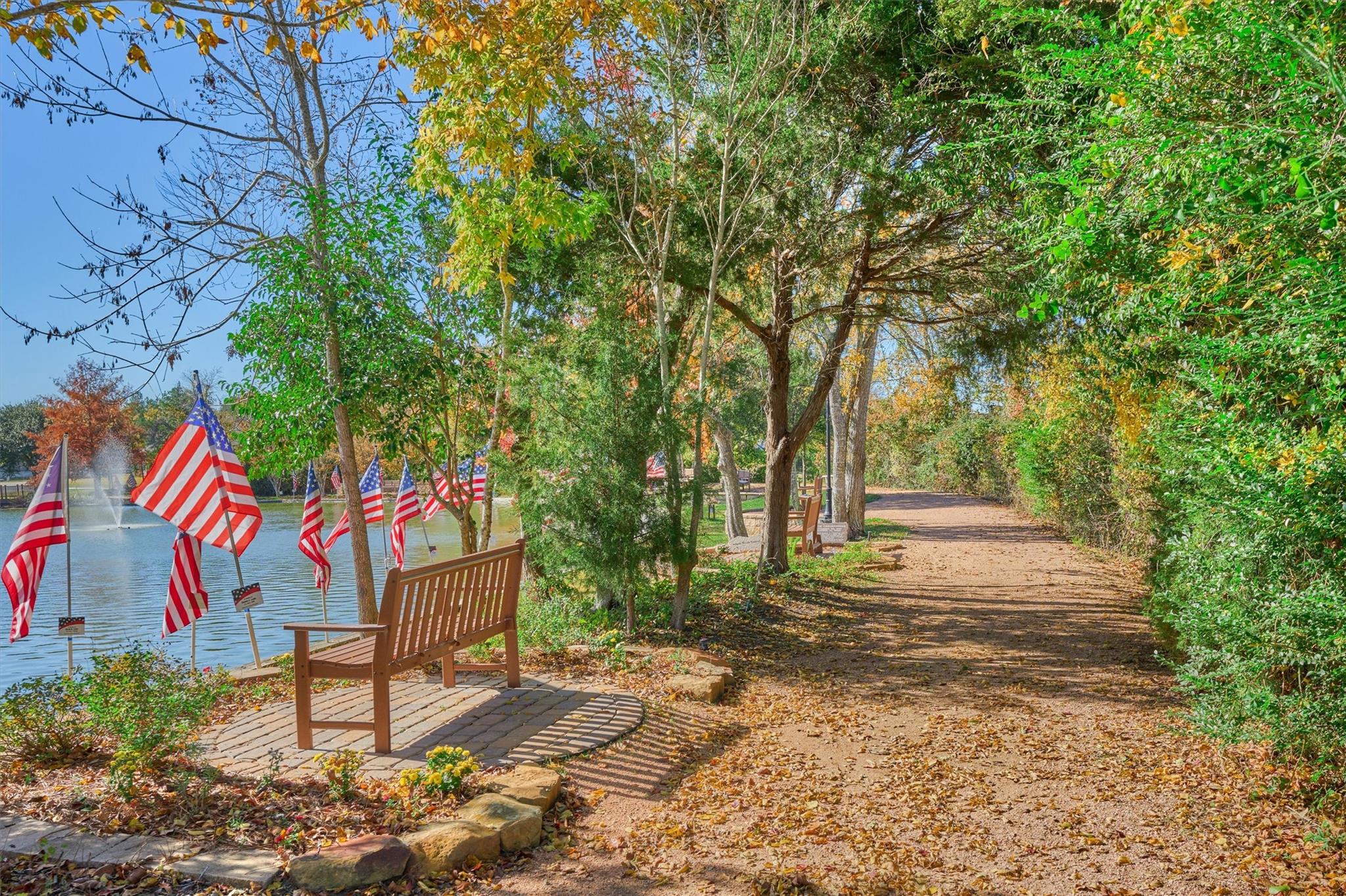 Pondside walking path with a fountain, wooden benches, and American flags beneath autumn trees.