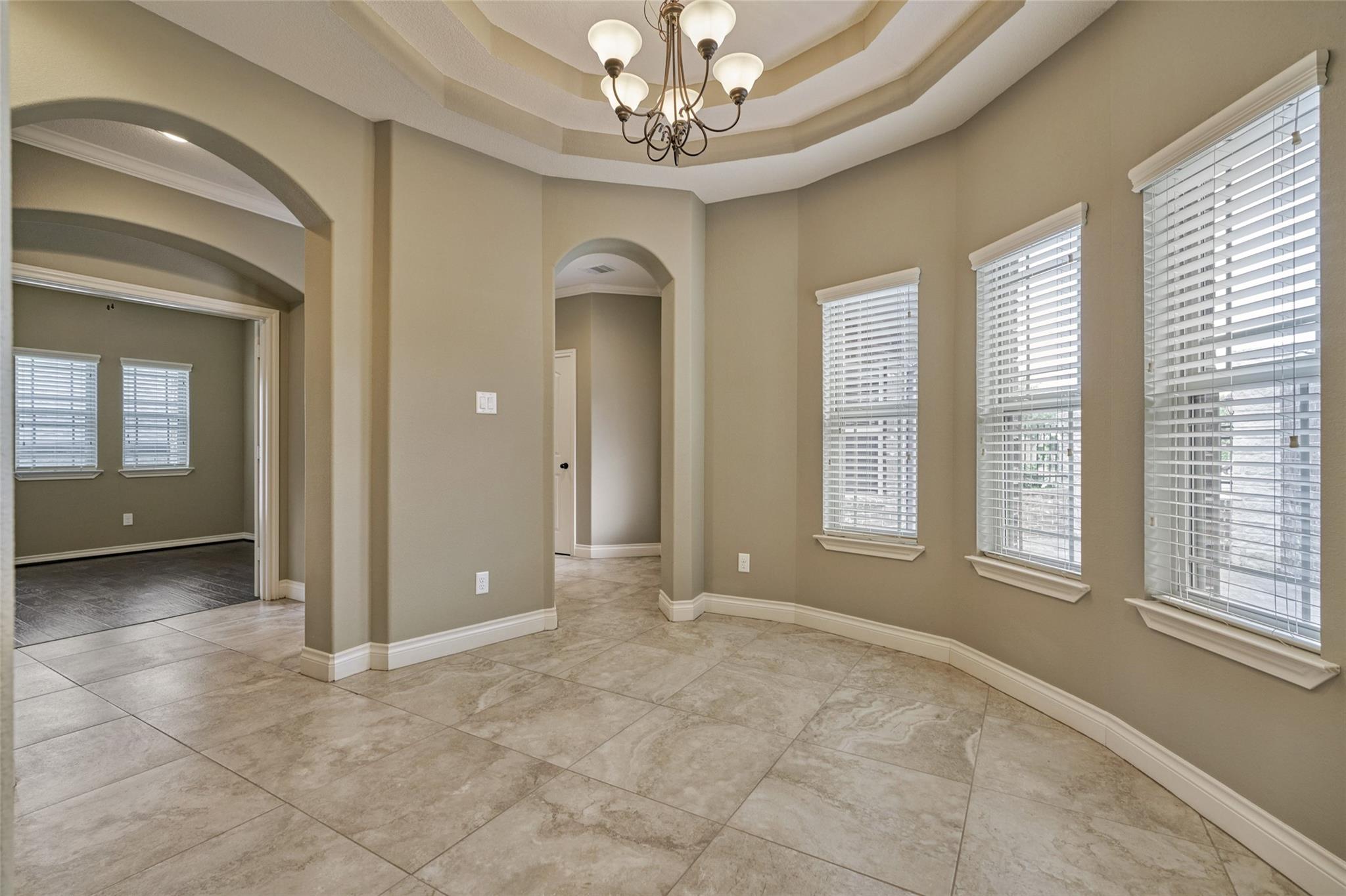 Dining area with a tiered ceiling, chandelier, tiled floor, and three windows with blinds.