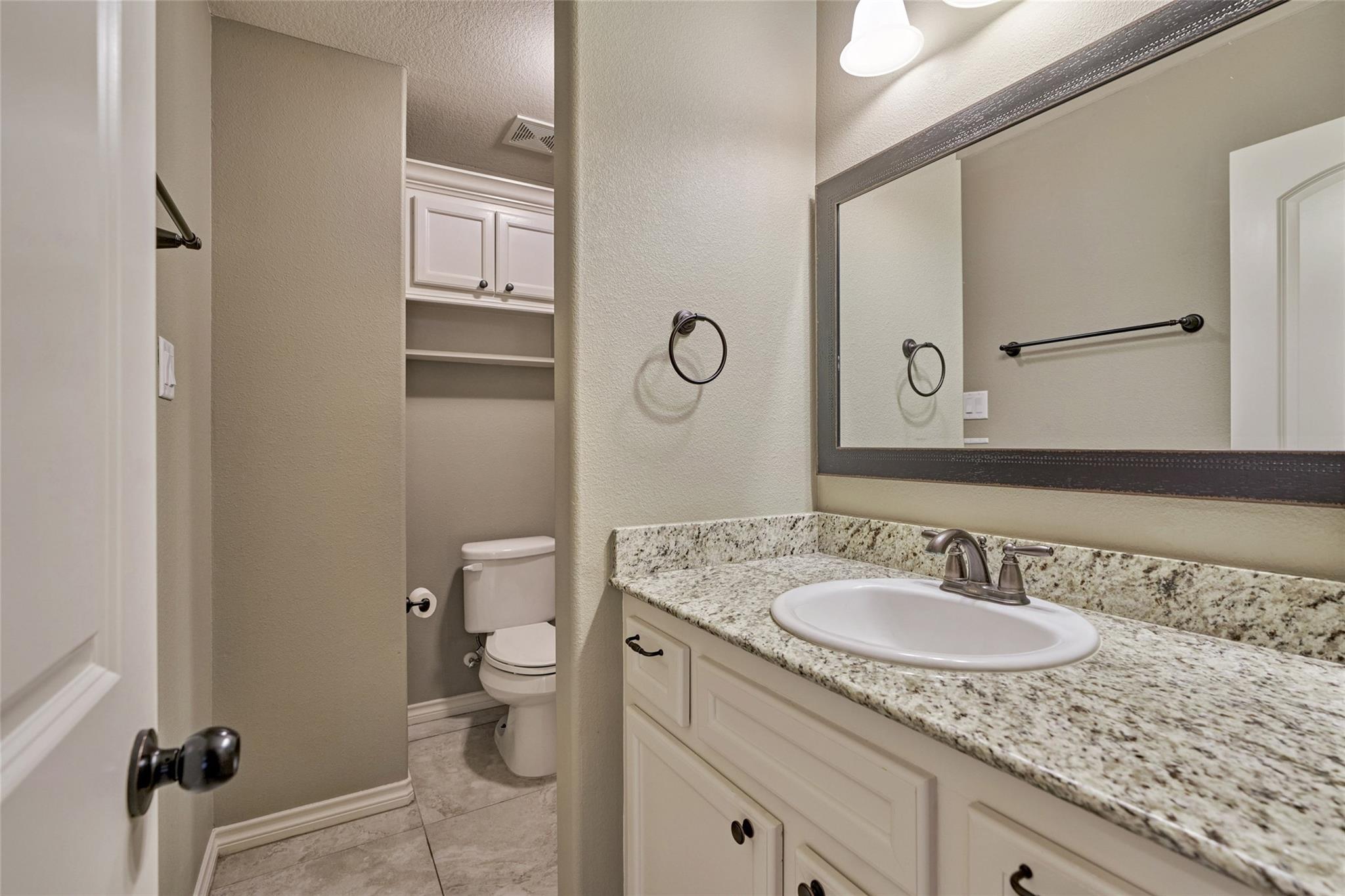 Bathroom vanity with granite countertop and large mirror beside a recessed toilet alcove.
