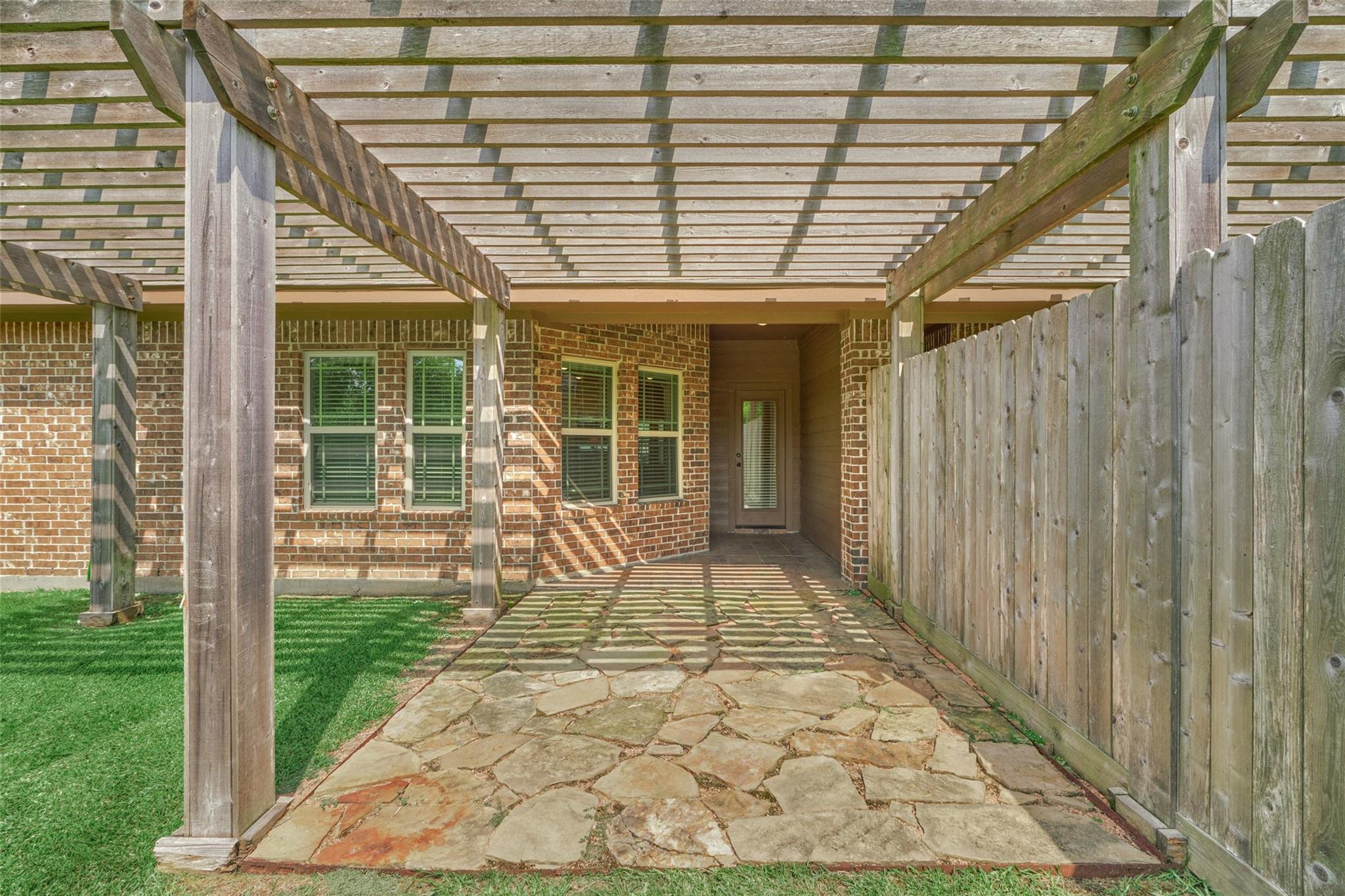 Covered stone patio with a wooden pergola, brick walls, and a tall privacy fence.
