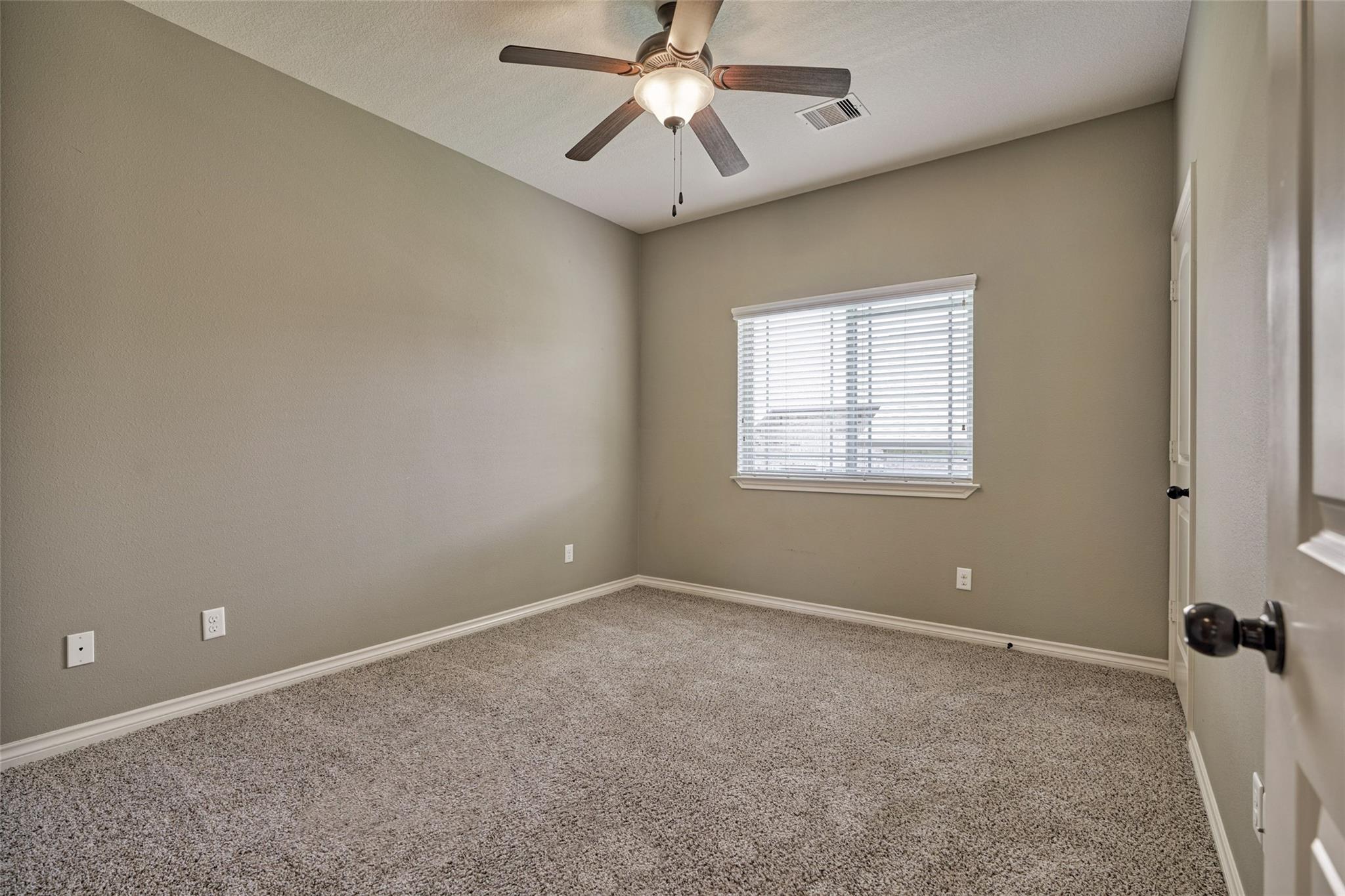 Empty carpeted bedroom with a ceiling fan and a window fitted with white blinds.