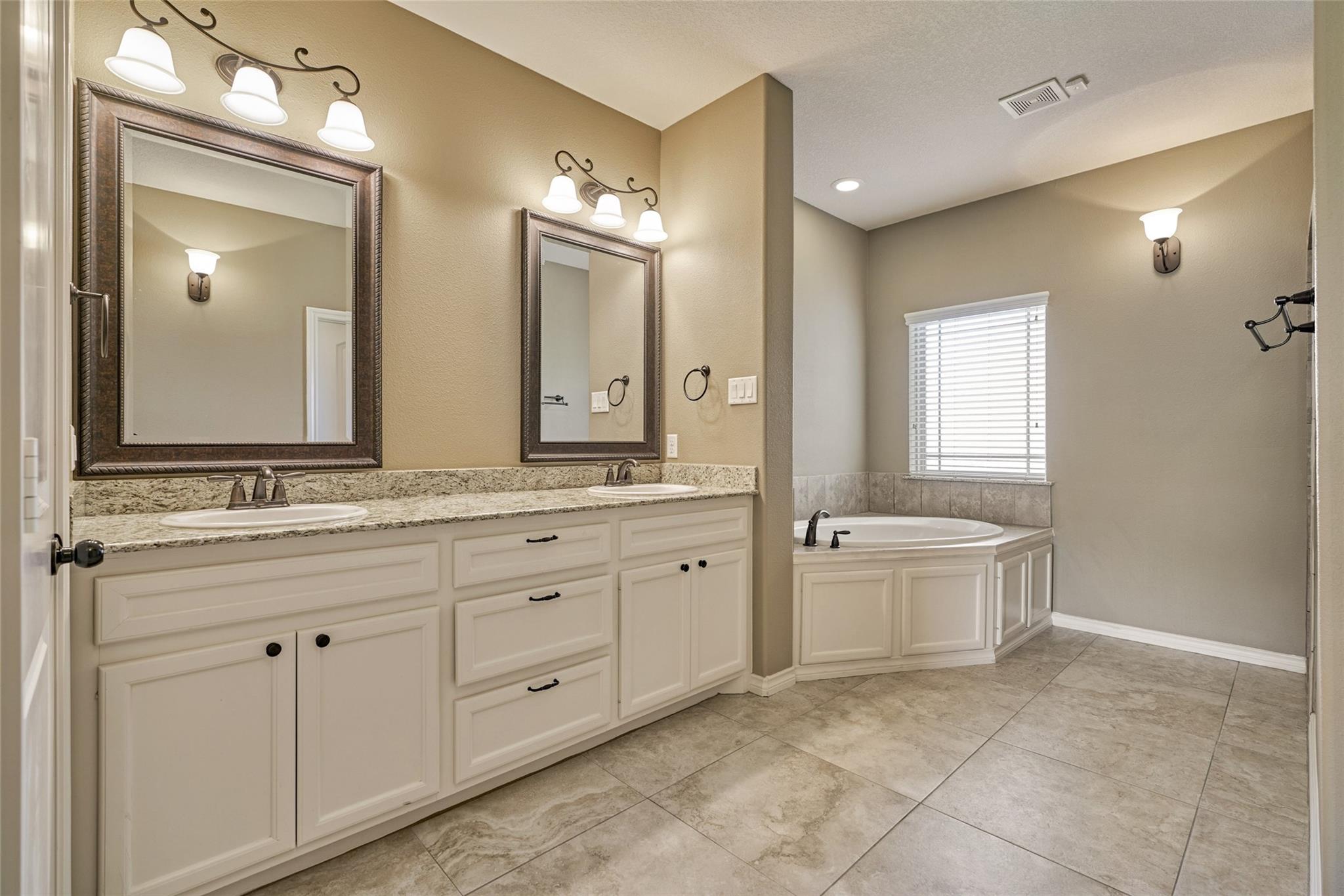 Bathroom with a double-sink granite vanity and corner soaking tub beneath a window.