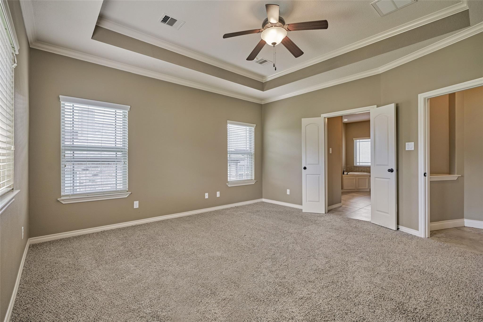 Empty carpeted bedroom with a tray ceiling, ceiling fan, and open doors to a bathroom and closet.