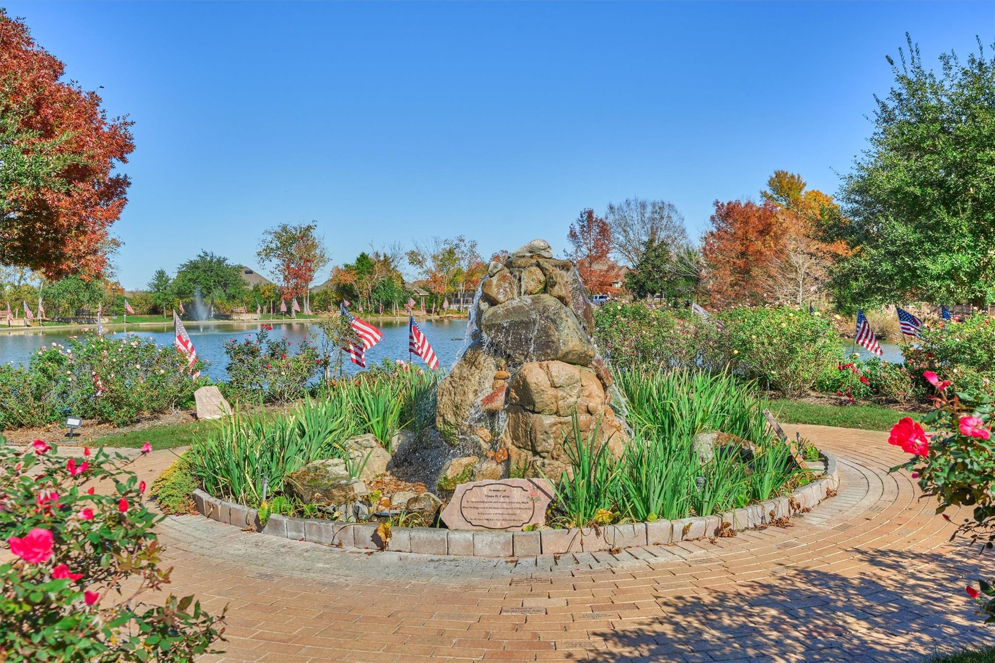 Community pond garden with a stone fountain, curved brick path, roses, and autumn foliage.