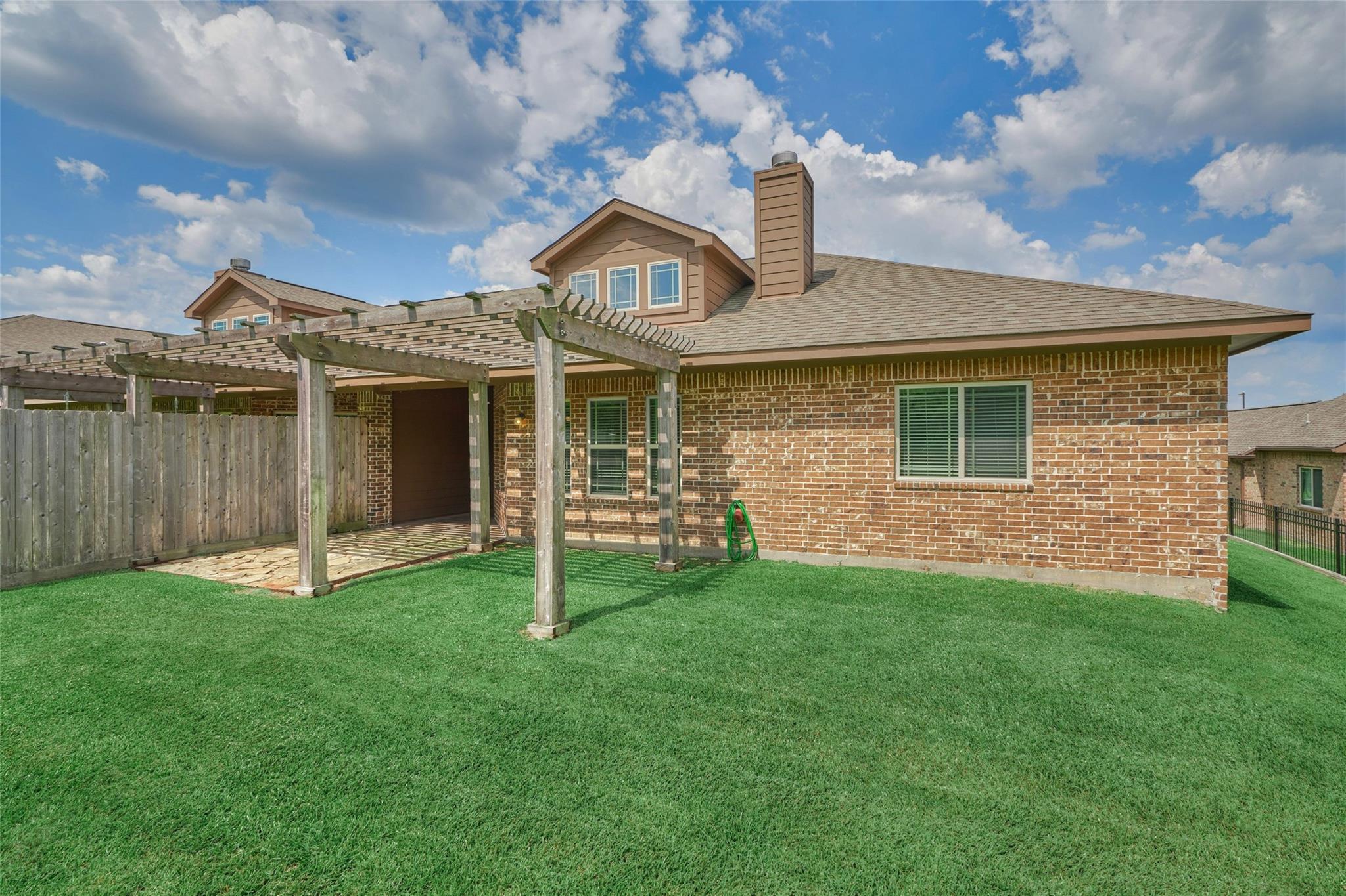 Brick home’s rear exterior with a wooden pergola, stone patio, and fenced lawn.