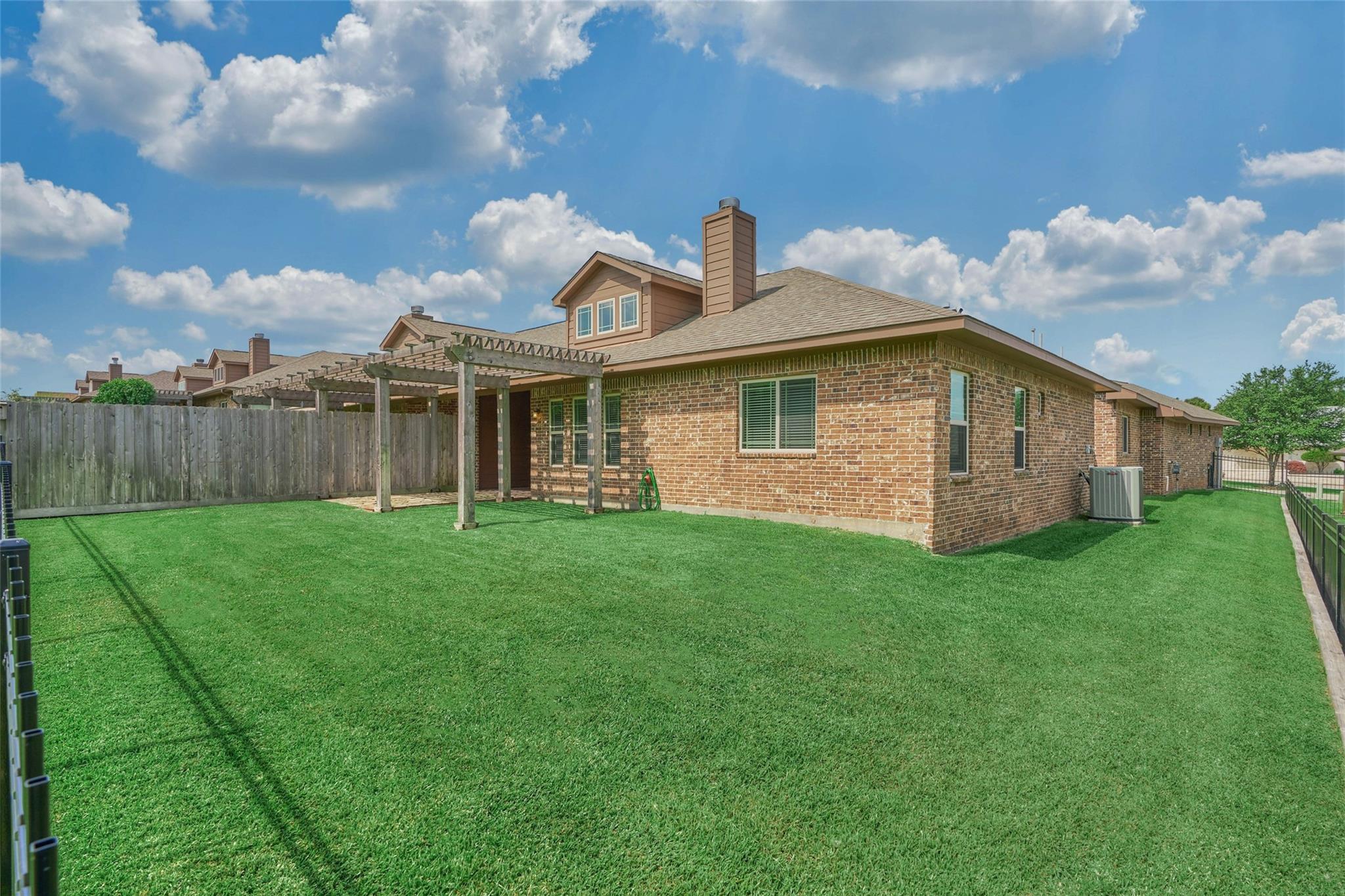 Brick home behind a broad fenced lawn, with a wooden pergola and neighboring houses.