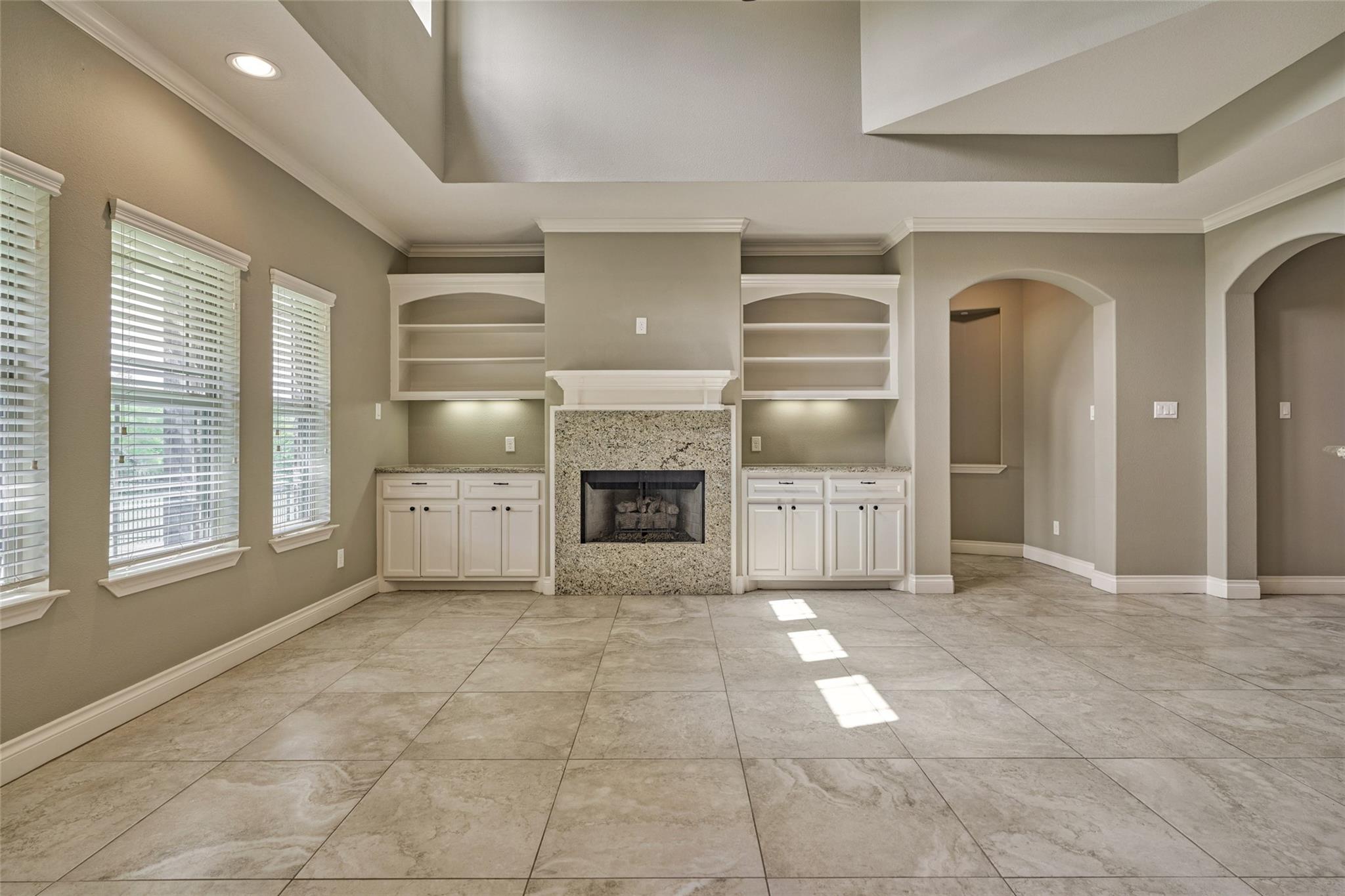 Empty living room with a stone fireplace, built-in shelving, tiled floors, and a tray ceiling.