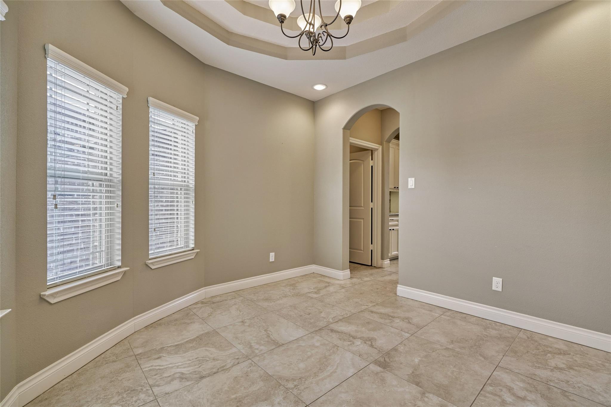 Empty tiled room with a tray ceiling, chandelier, two windows, and an arched doorway.