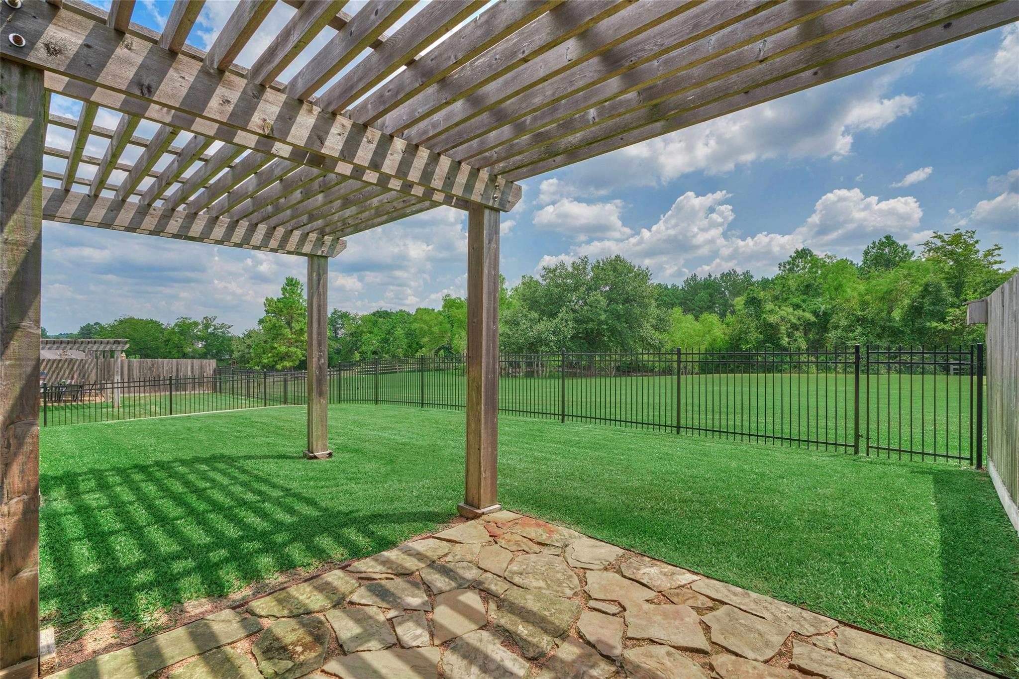 Backyard patio beneath a wooden pergola, overlooking a fenced green lawn and mature trees.