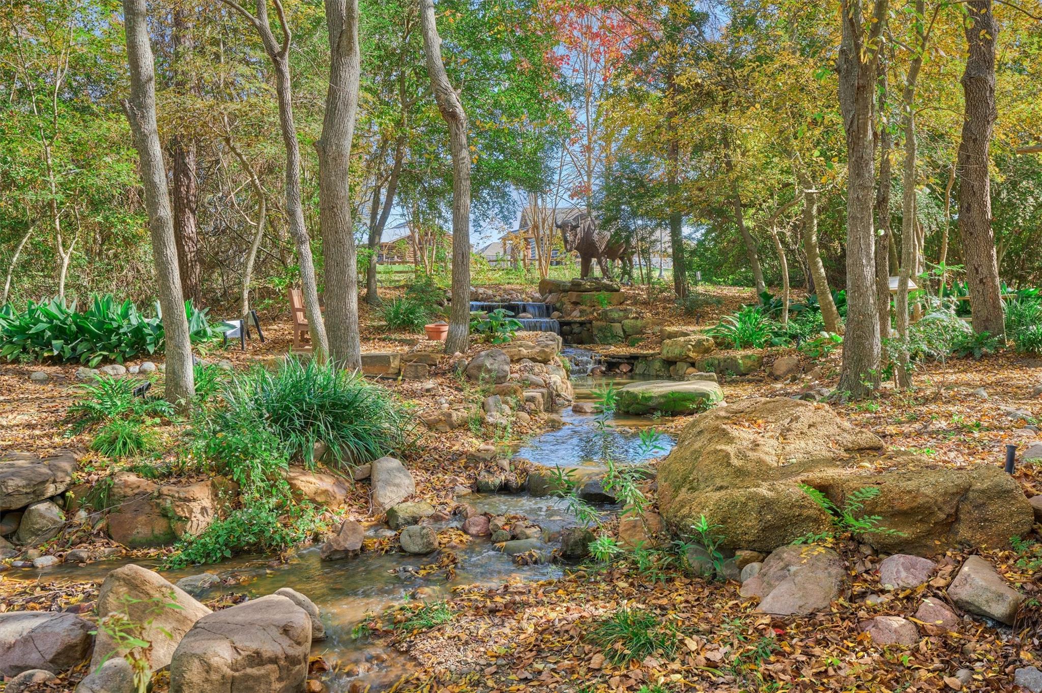 Wooded garden with a rock-lined creek, cascading waterfall, and animal sculpture.