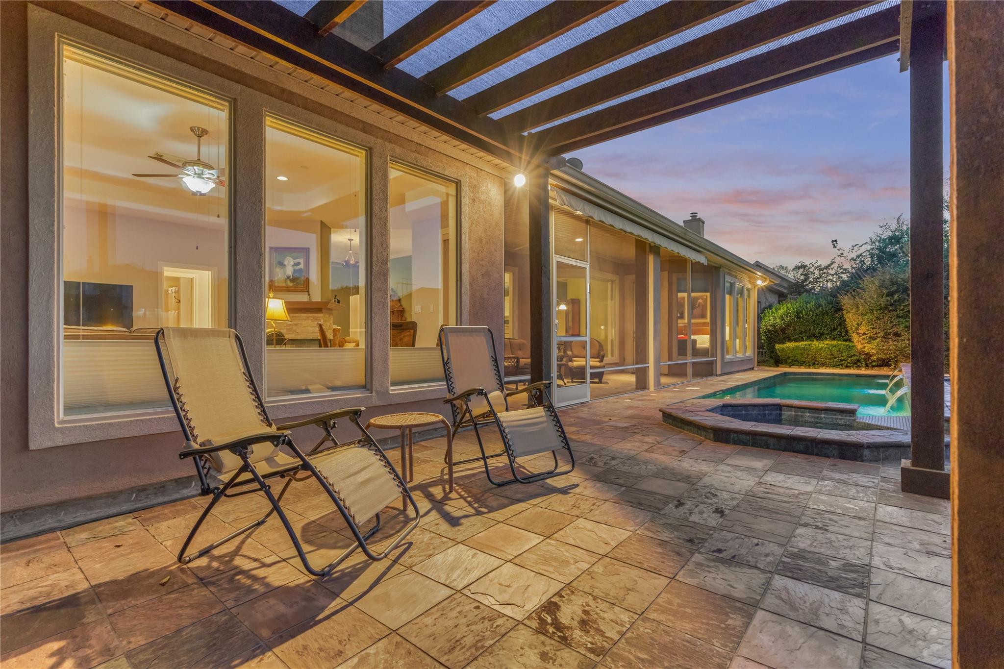 Covered patio with lounge chairs beside a pool and raised spa at dusk.
