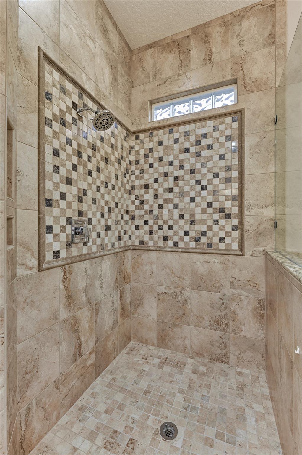 Walk-in shower with a beige stone tile floor and checkerboard mosaic walls beneath a glass-block window.