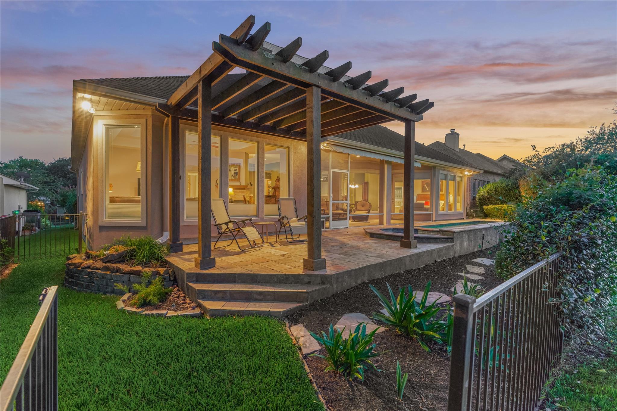 Home’s rear patio at dusk, with a timber pergola, outdoor chairs, landscaped garden beds, and a small pool.