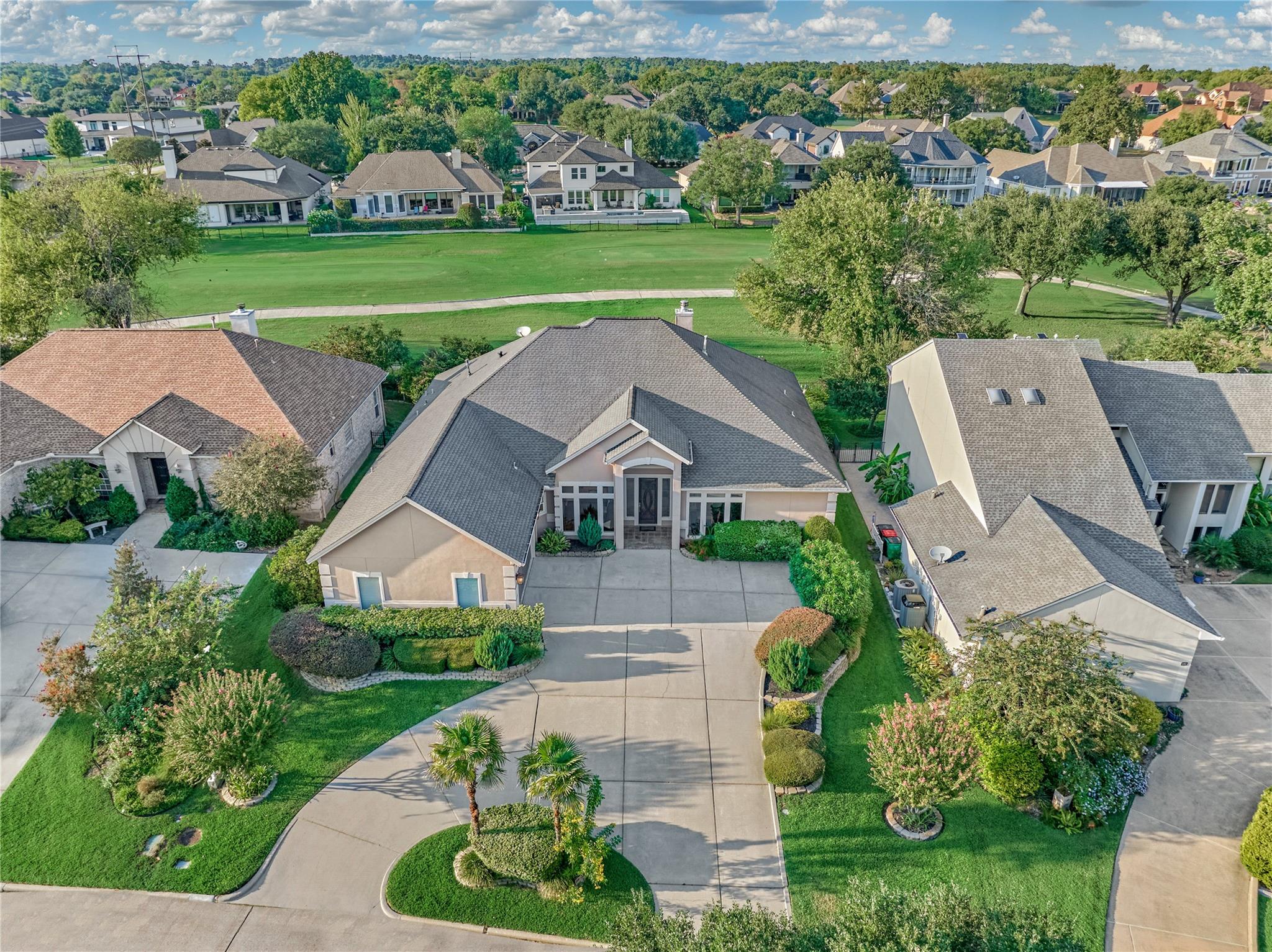 Aerial view of a landscaped home with a curved driveway and green open space behind it.