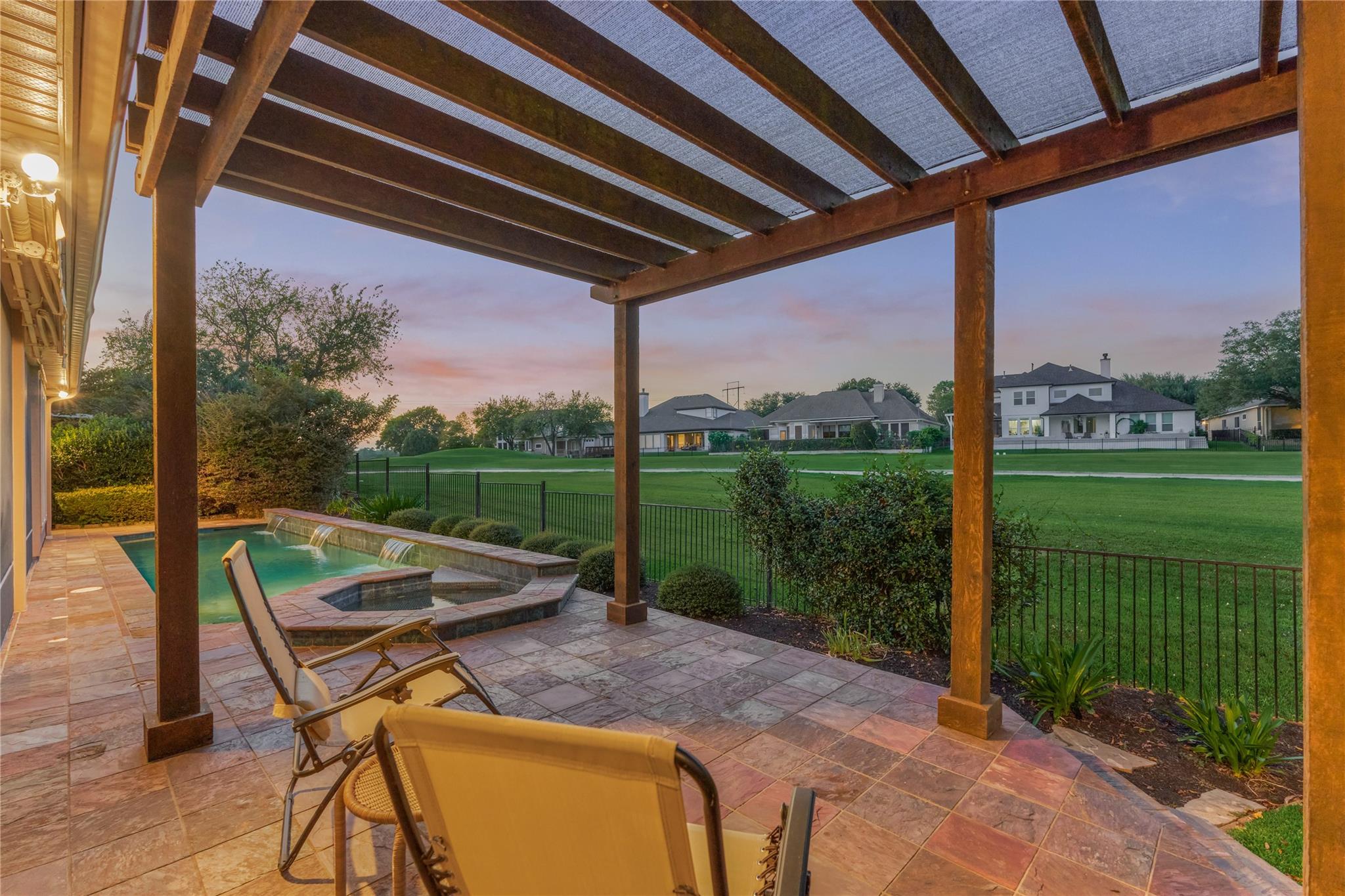 Covered patio with a timber pergola overlooks a pool, spa, and green lawn at dusk.