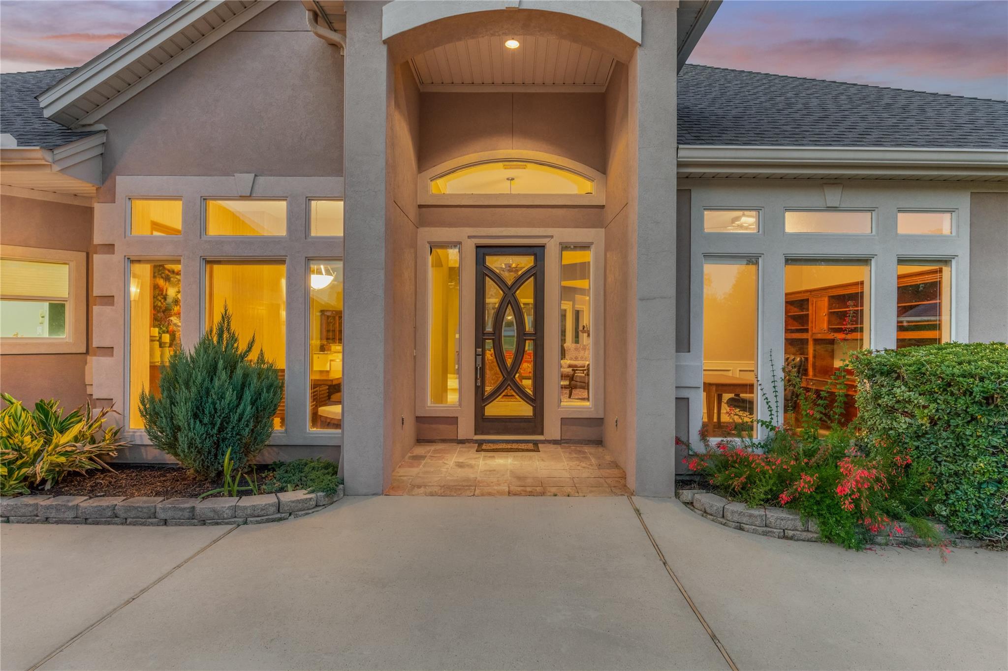 Gray stucco home with a tall covered entry, decorative glass door, lit windows, and landscaped front beds.