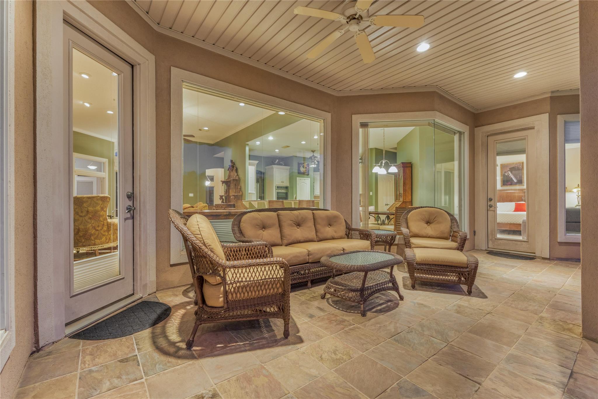 Covered patio with wicker seating, tiled floor, ceiling fan, and large windows into the home.