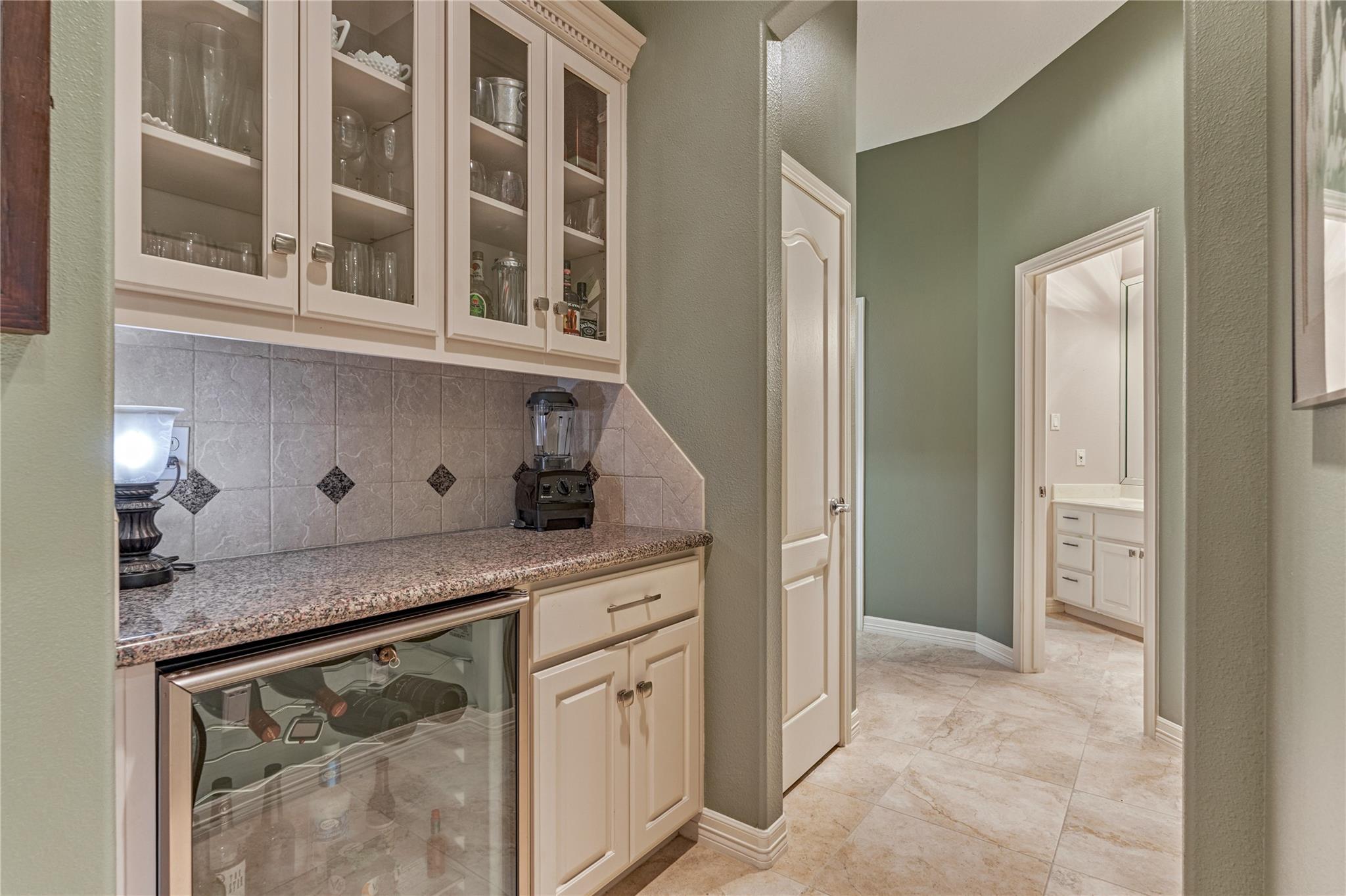 Wet bar with glass-front cabinets, granite counter, and beverage refrigerator beside a tiled hallway.