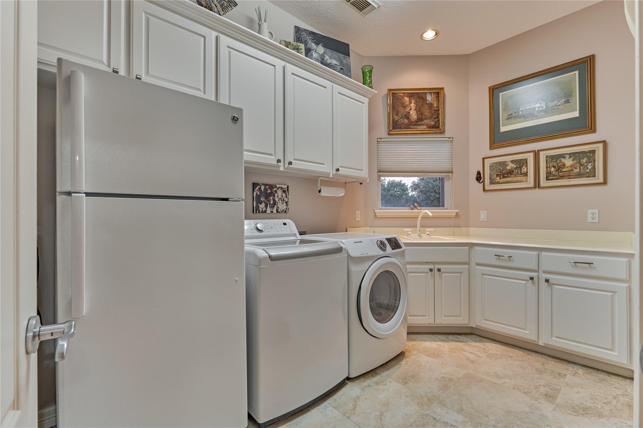 Laundry room with white cabinets, washer and dryer, refrigerator, and a sink beneath a window.