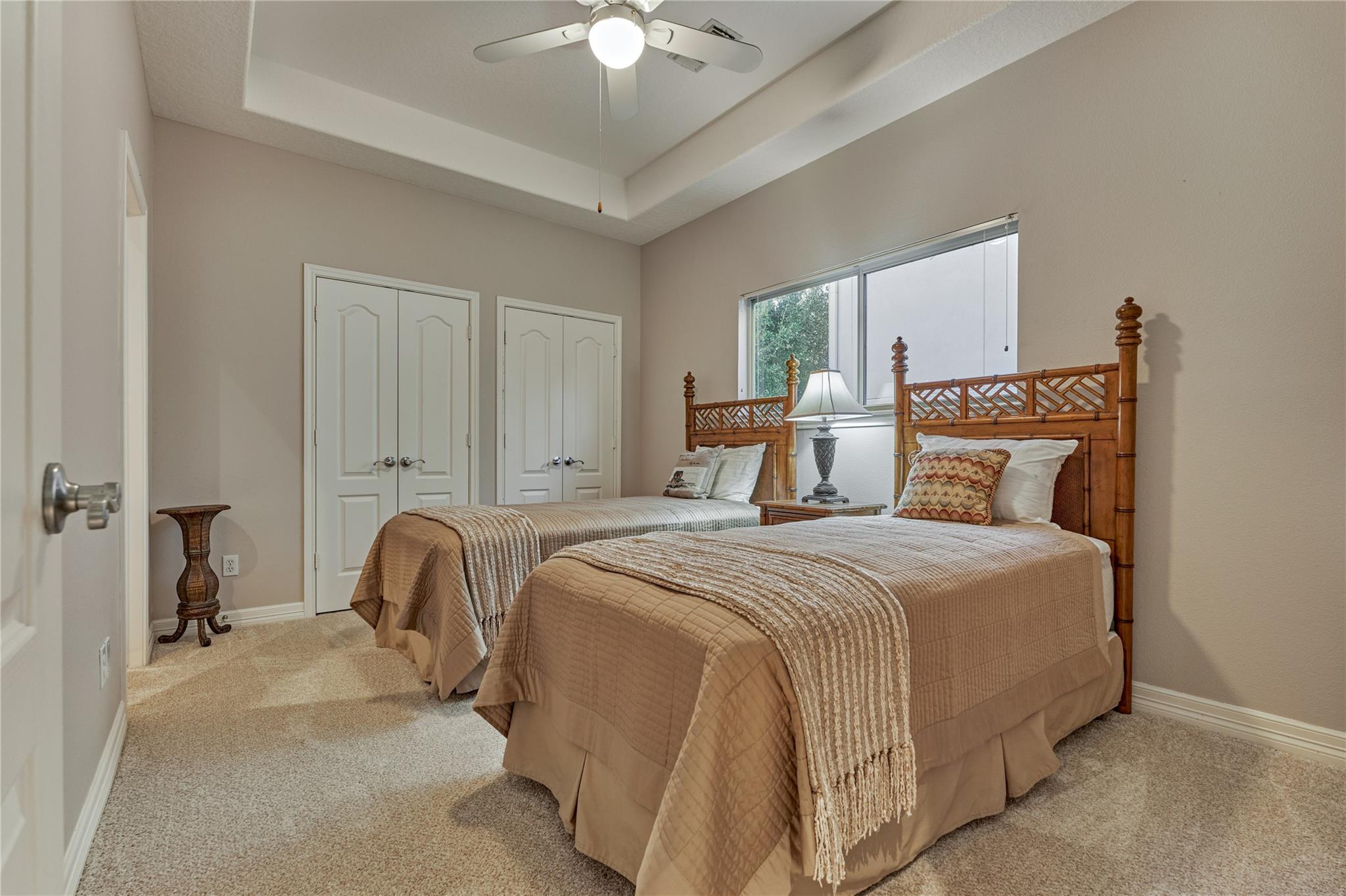 Bedroom with two wooden beds, beige bedding, carpet, and a tray ceiling with ceiling fan.