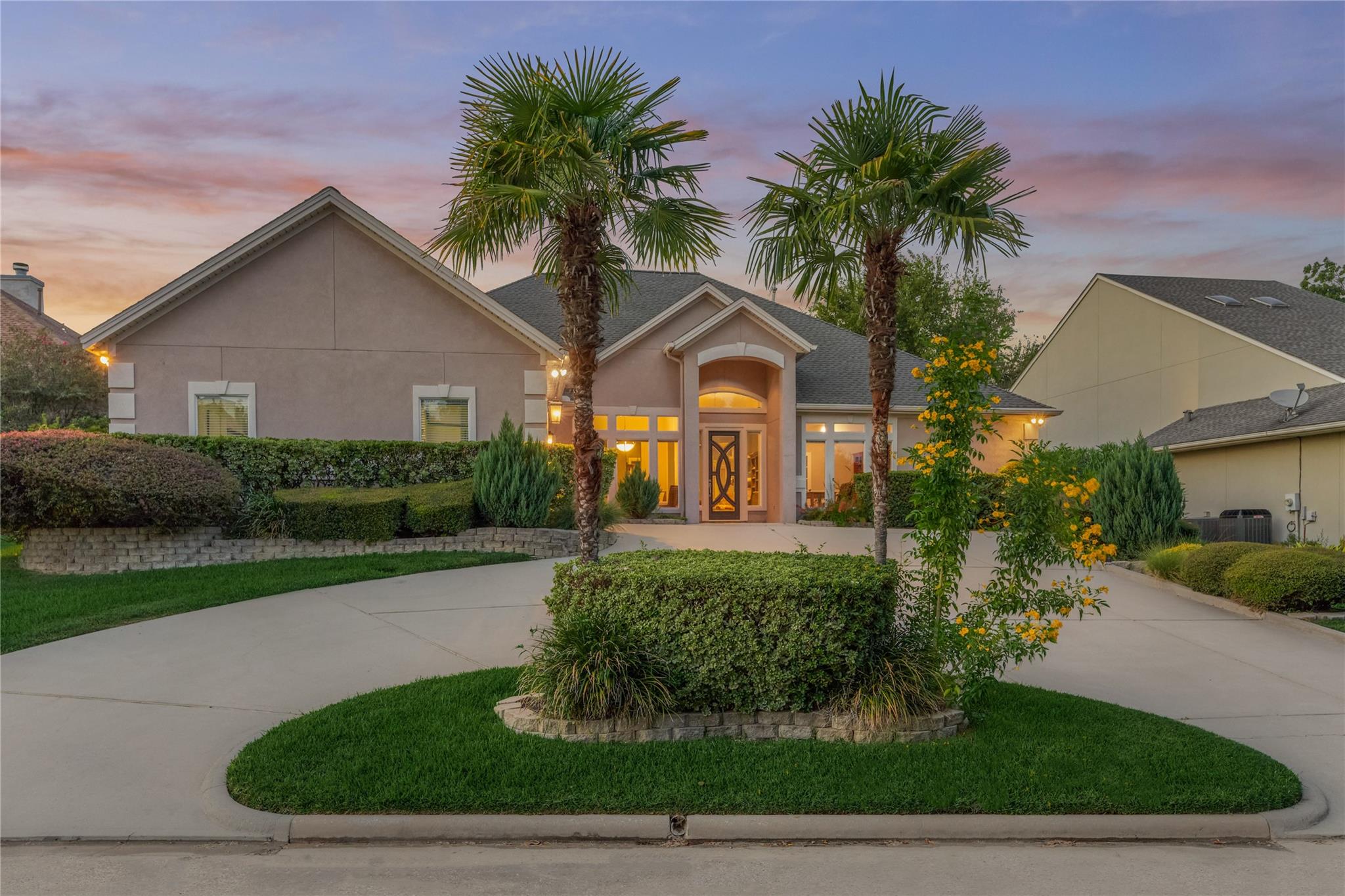 Stucco home at dusk with a central arched entry, palm trees, and a curved driveway.