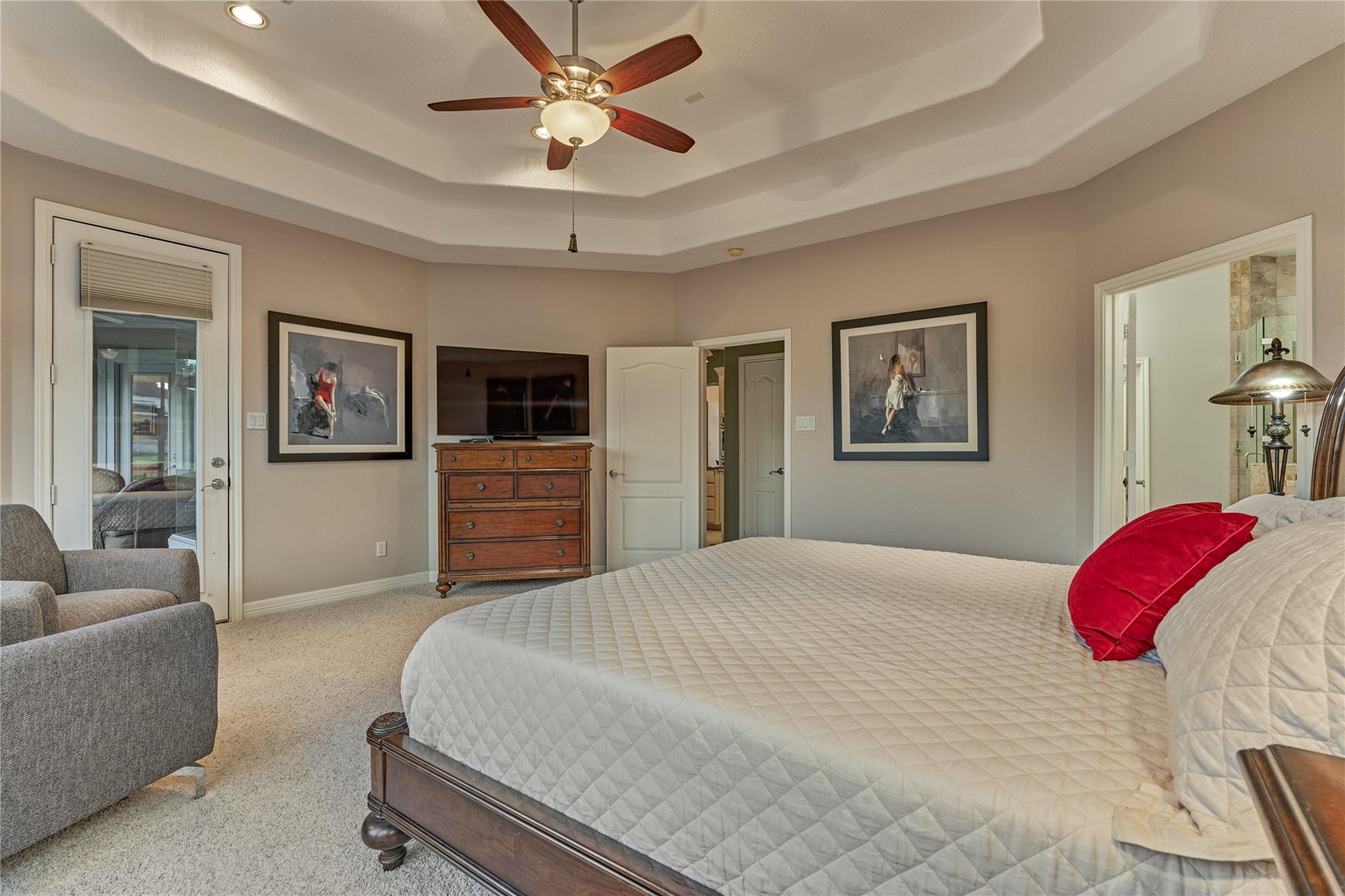 Furnished bedroom with a large bed, tray ceiling, ceiling fan, and television on a wooden dresser.