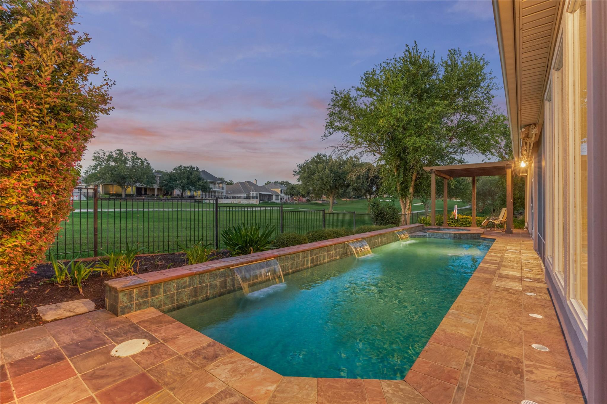 Backyard pool with cascading water features, tiled patio, and covered pergola at dusk.