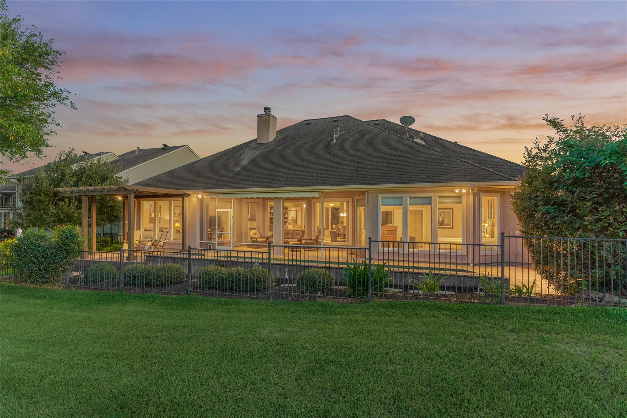 Home’s rear exterior at dusk, with illuminated windows, a covered patio, pergola, and fenced lawn.