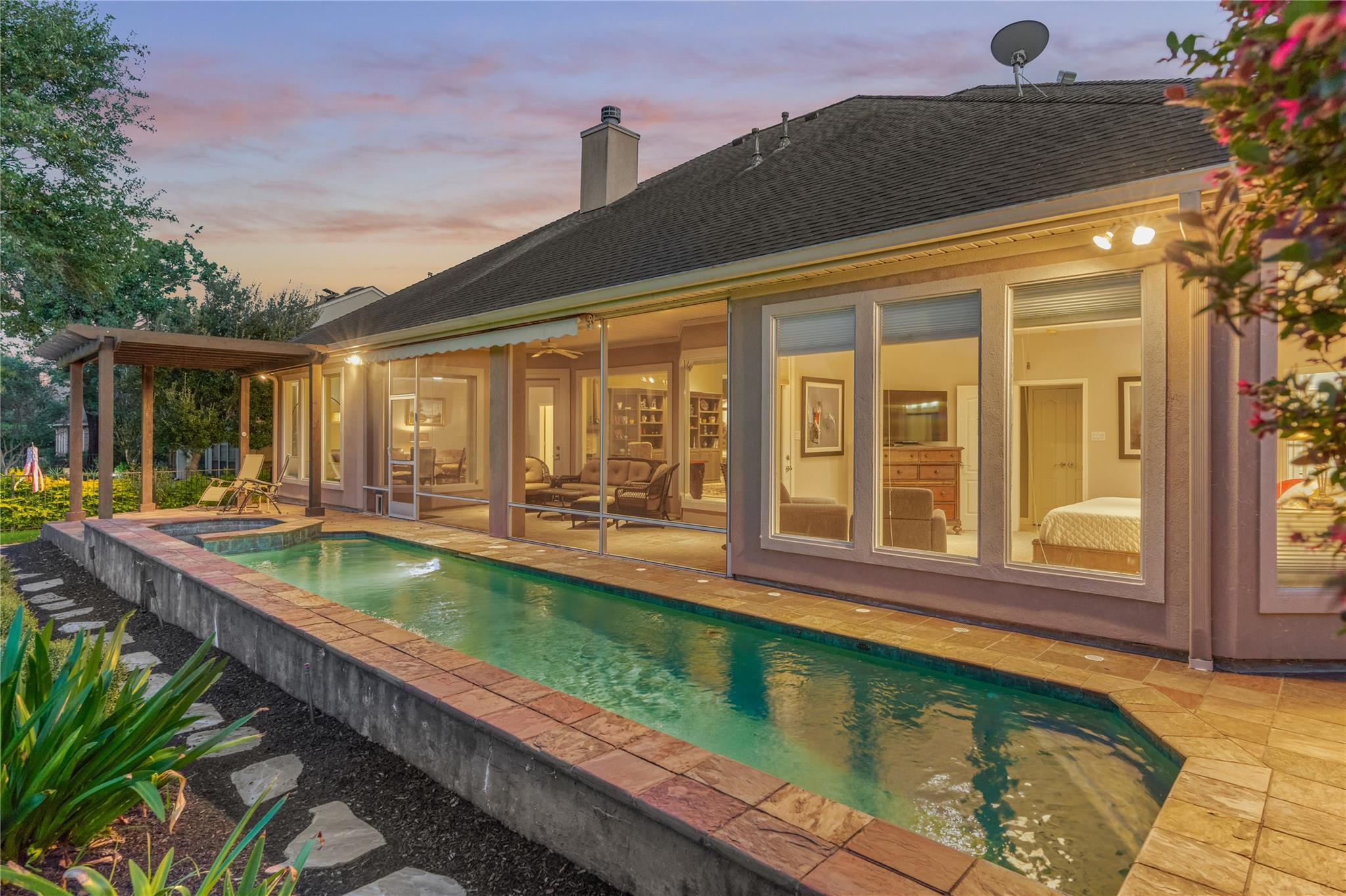 Home’s rear exterior at dusk, with a long pool, raised spa, and covered pergola patio.