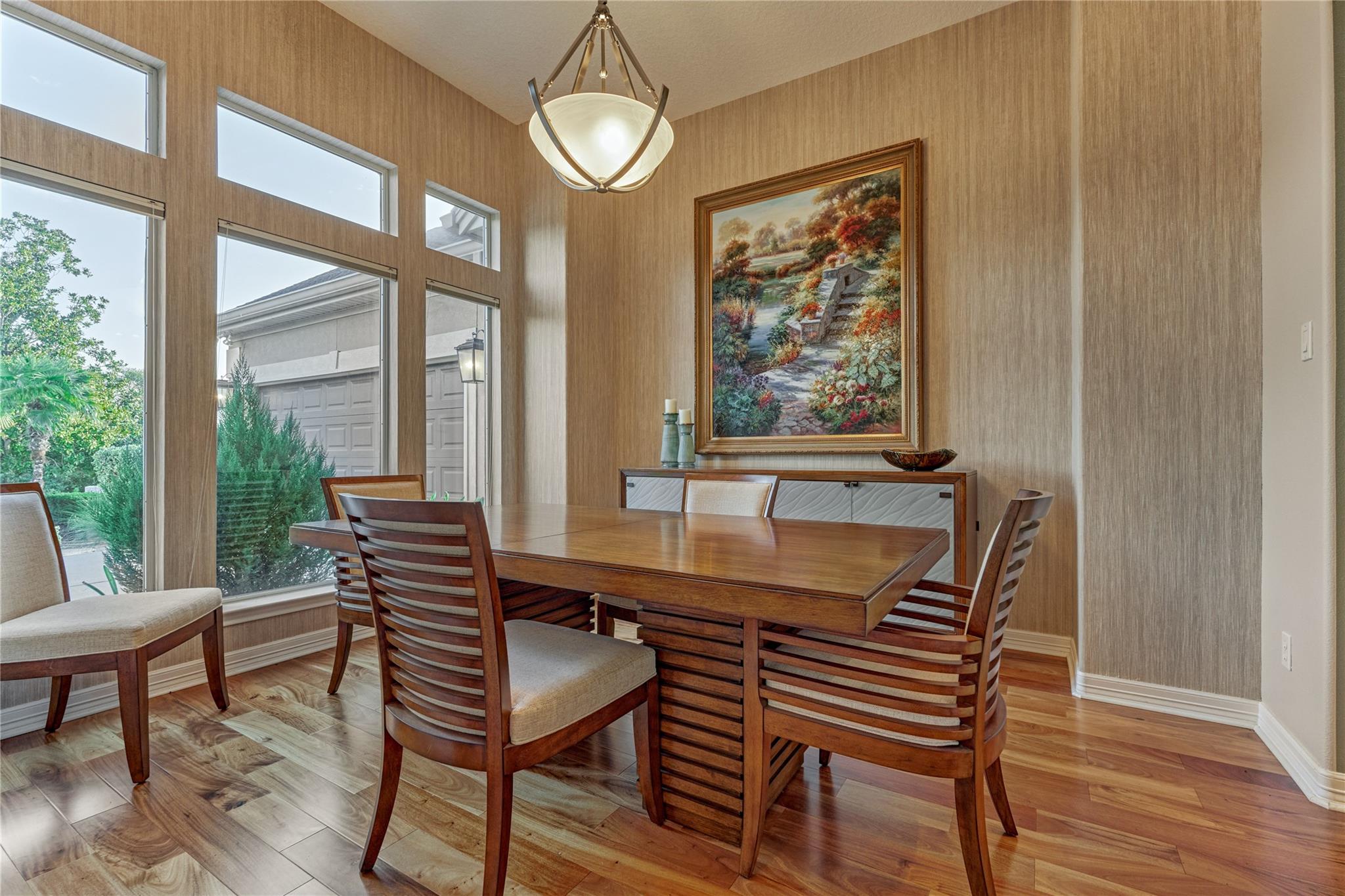Dining room with a wooden table, tall windows overlooking greenery, and hardwood floors.