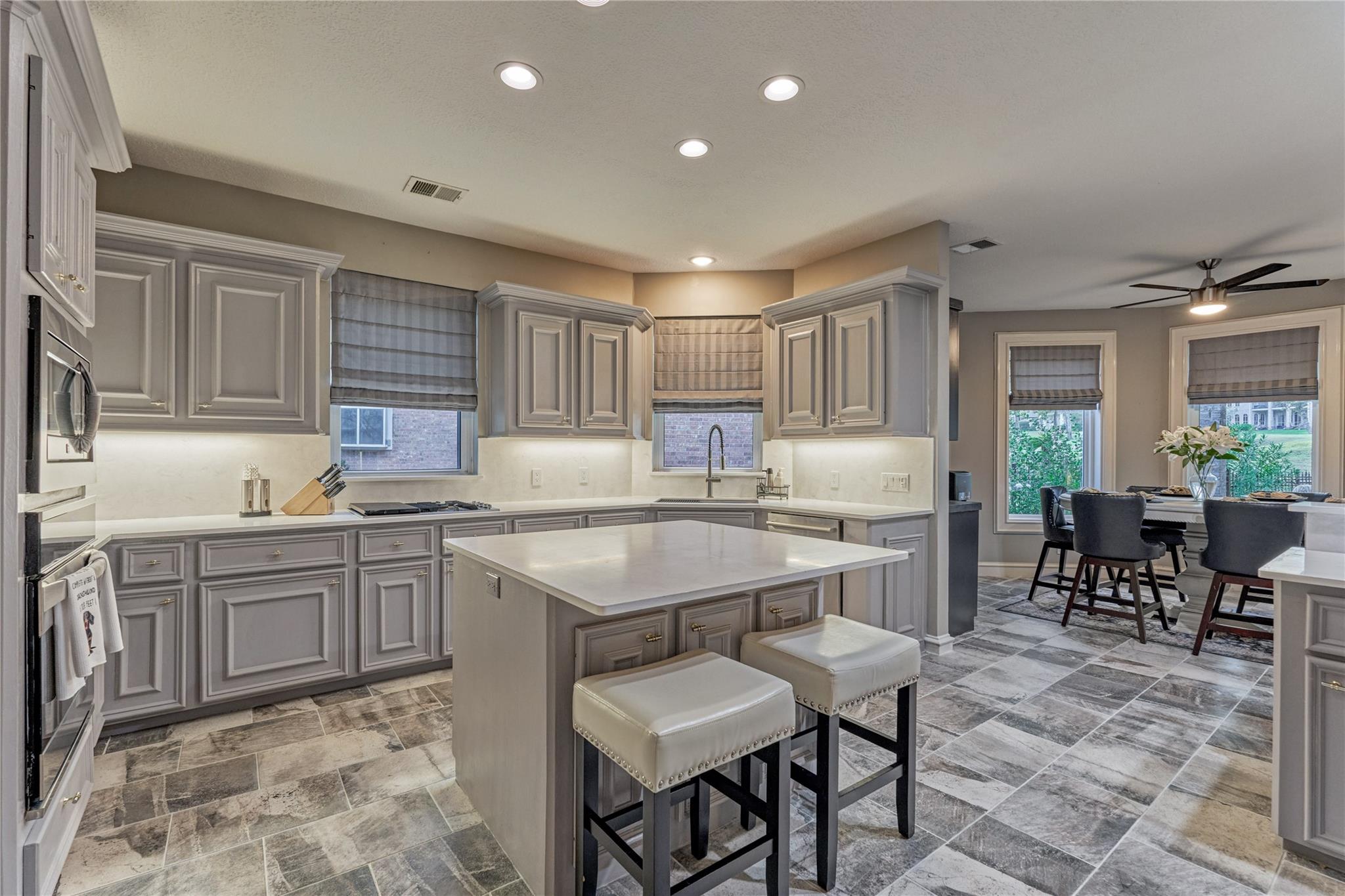 Kitchen with gray cabinetry, a large island and stools, opening to a windowed dining area.