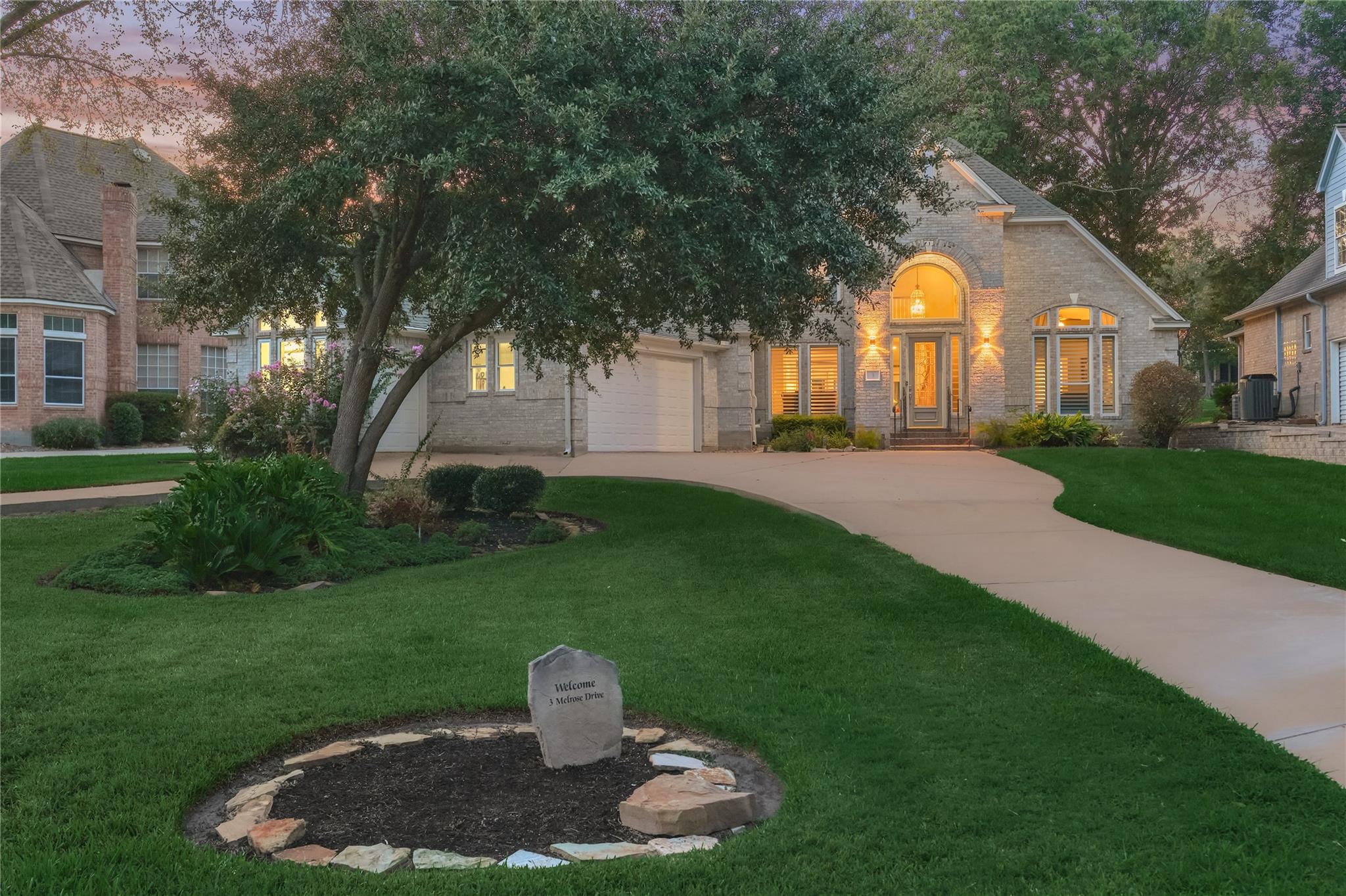 Light-brick home with a glowing arched entry, curved driveway, and landscaped lawn at dusk.
