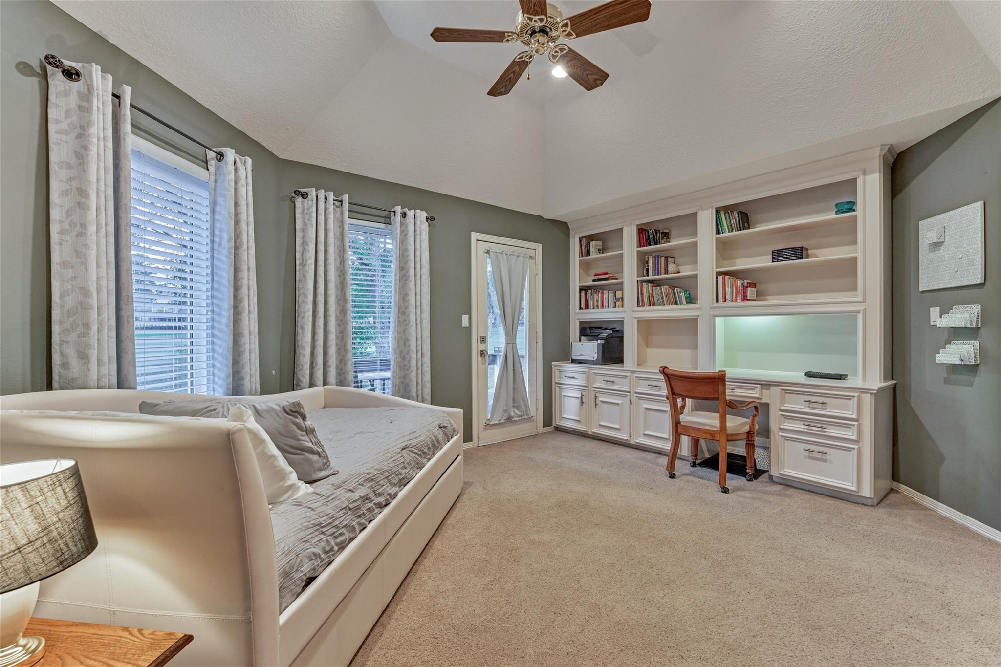 Furnished room with built-in desk and bookcases, a white daybed, and curtained windows.