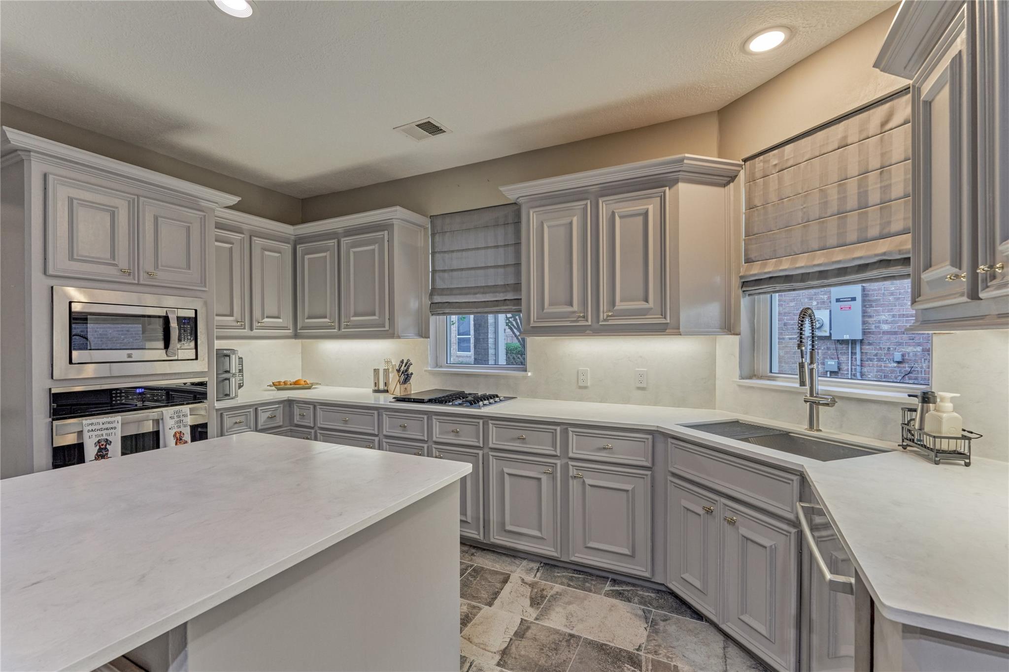 Kitchen with gray cabinetry, a white island, gas cooktop, and stainless sink beneath shaded windows.
