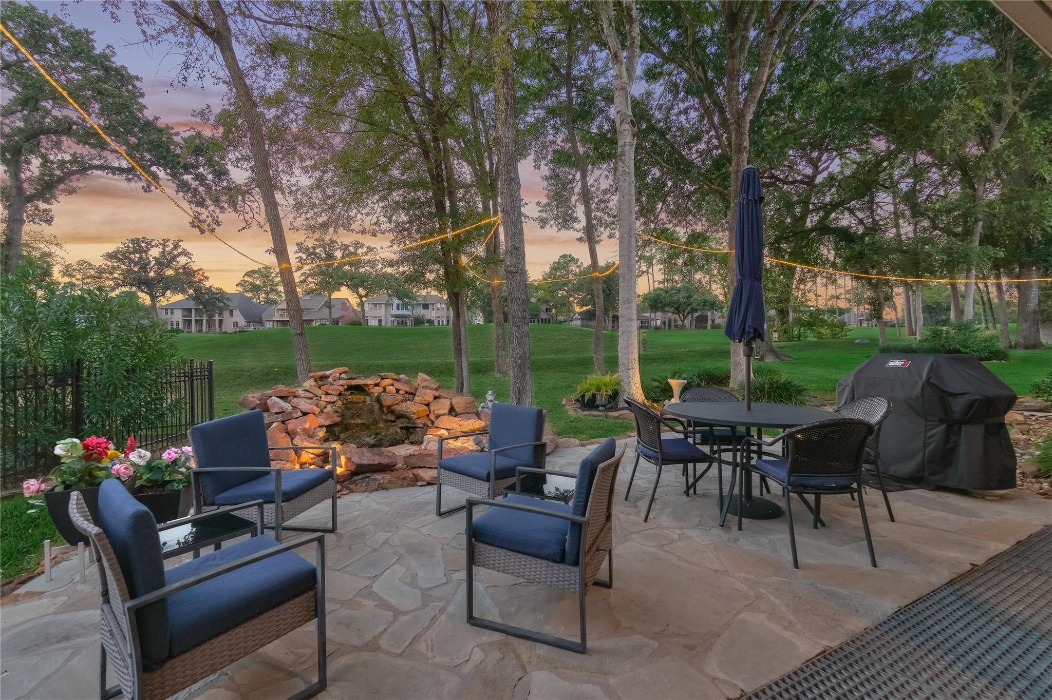 Stone patio with cushioned seating, a rock water feature, and string lights among mature trees.