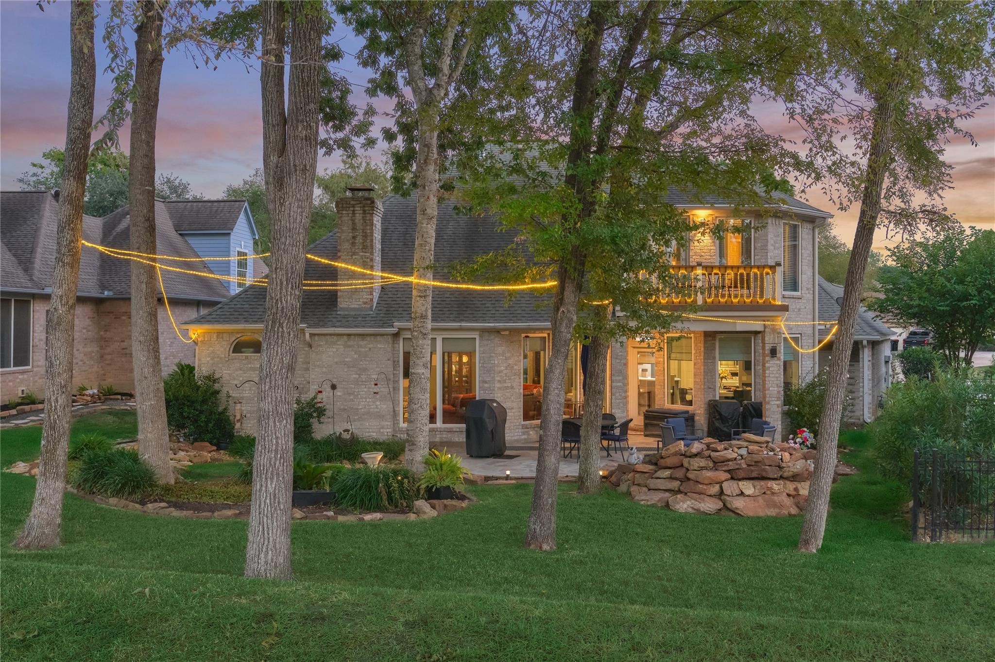Two-story brick home at dusk, with a tree-framed patio, illuminated balcony, and glowing string lights.