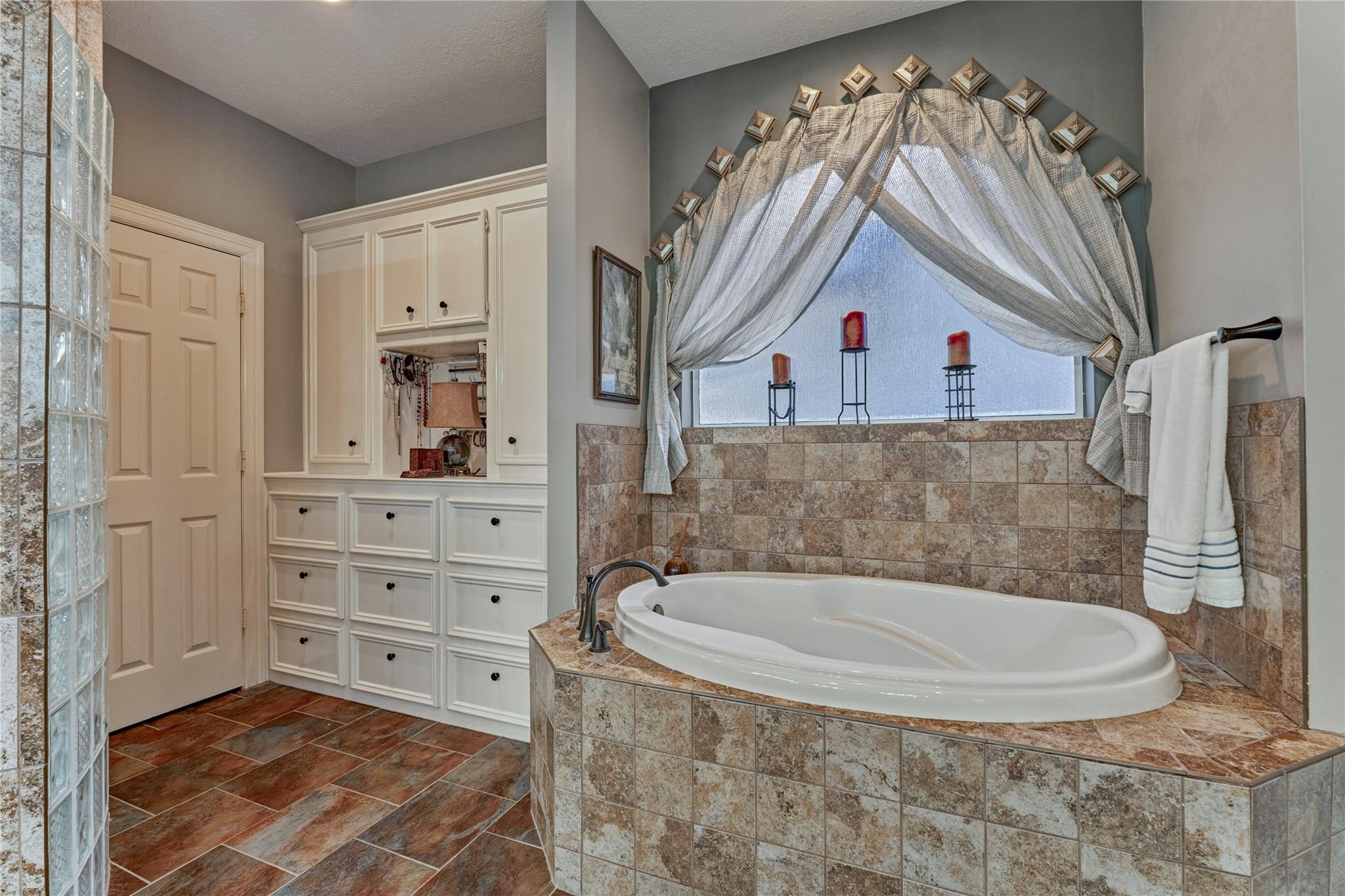 Bathroom with a large oval soaking tub, tiled surround, curtained window, and white built-in cabinets.