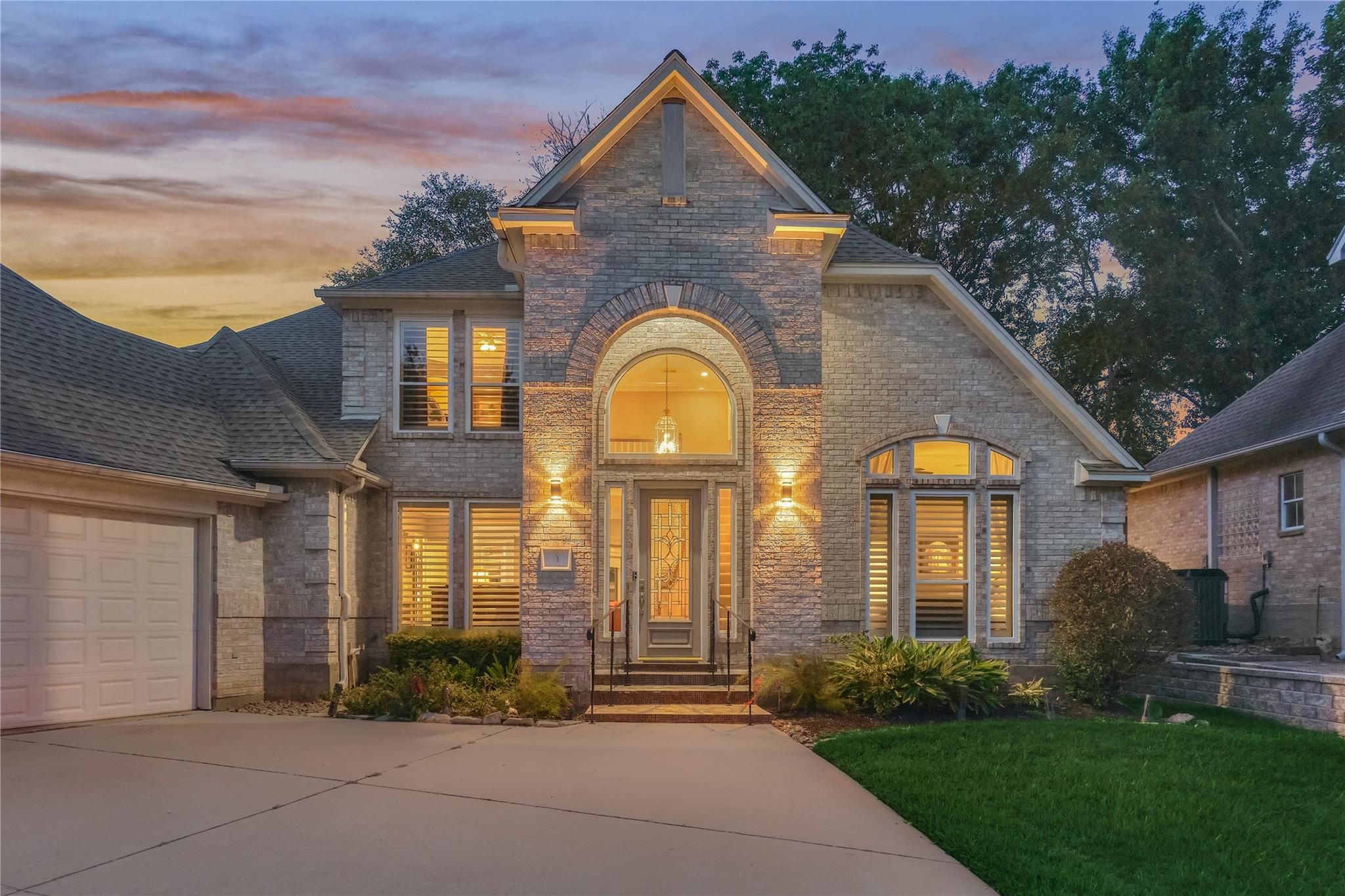Two-story stone house at dusk with an arched entry, illuminated windows, and an attached garage.